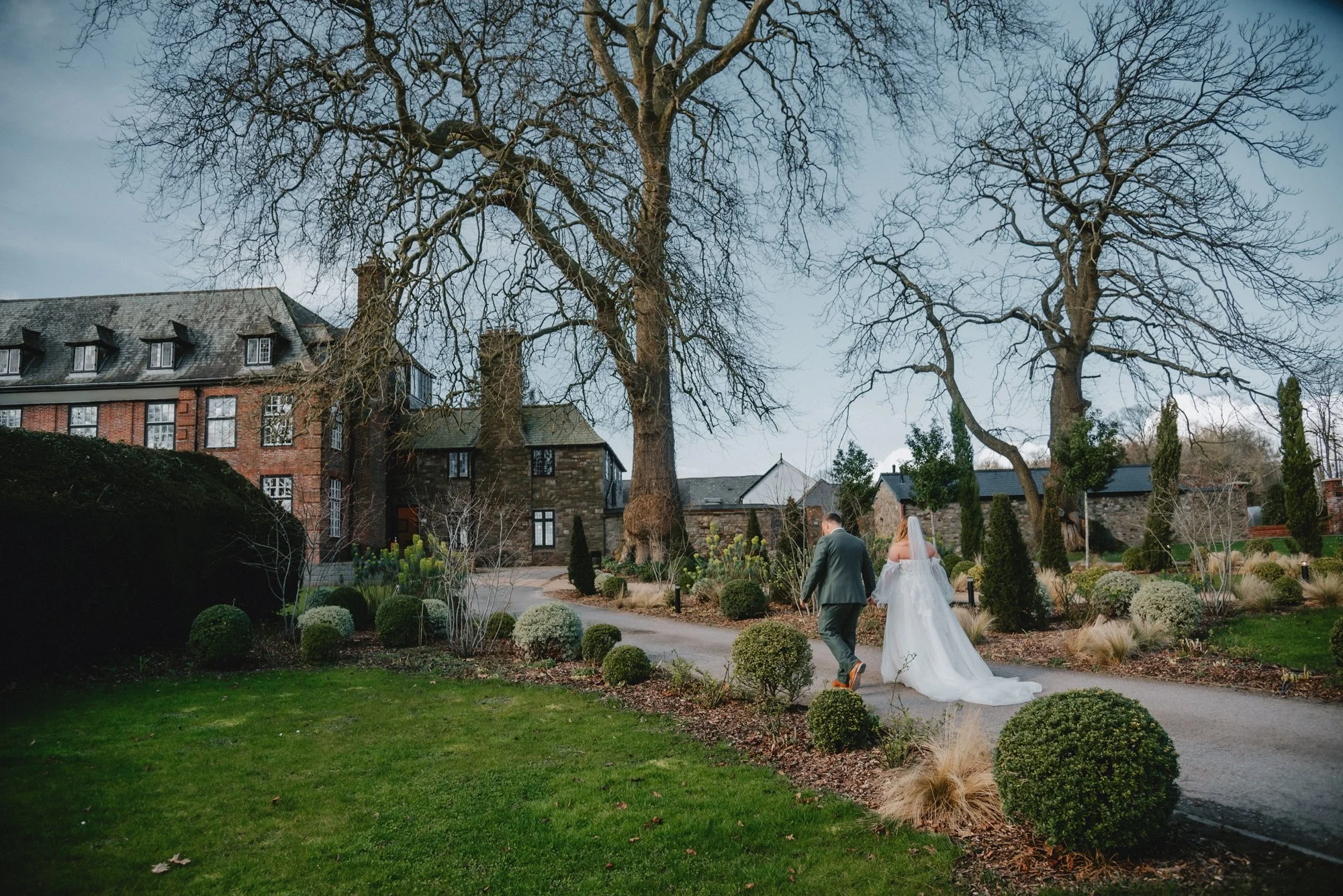 A bride and groom walk hand in hand along a garden path with manicured shrubs and large leafless trees, with historic buildings in the background under an overcast sky.