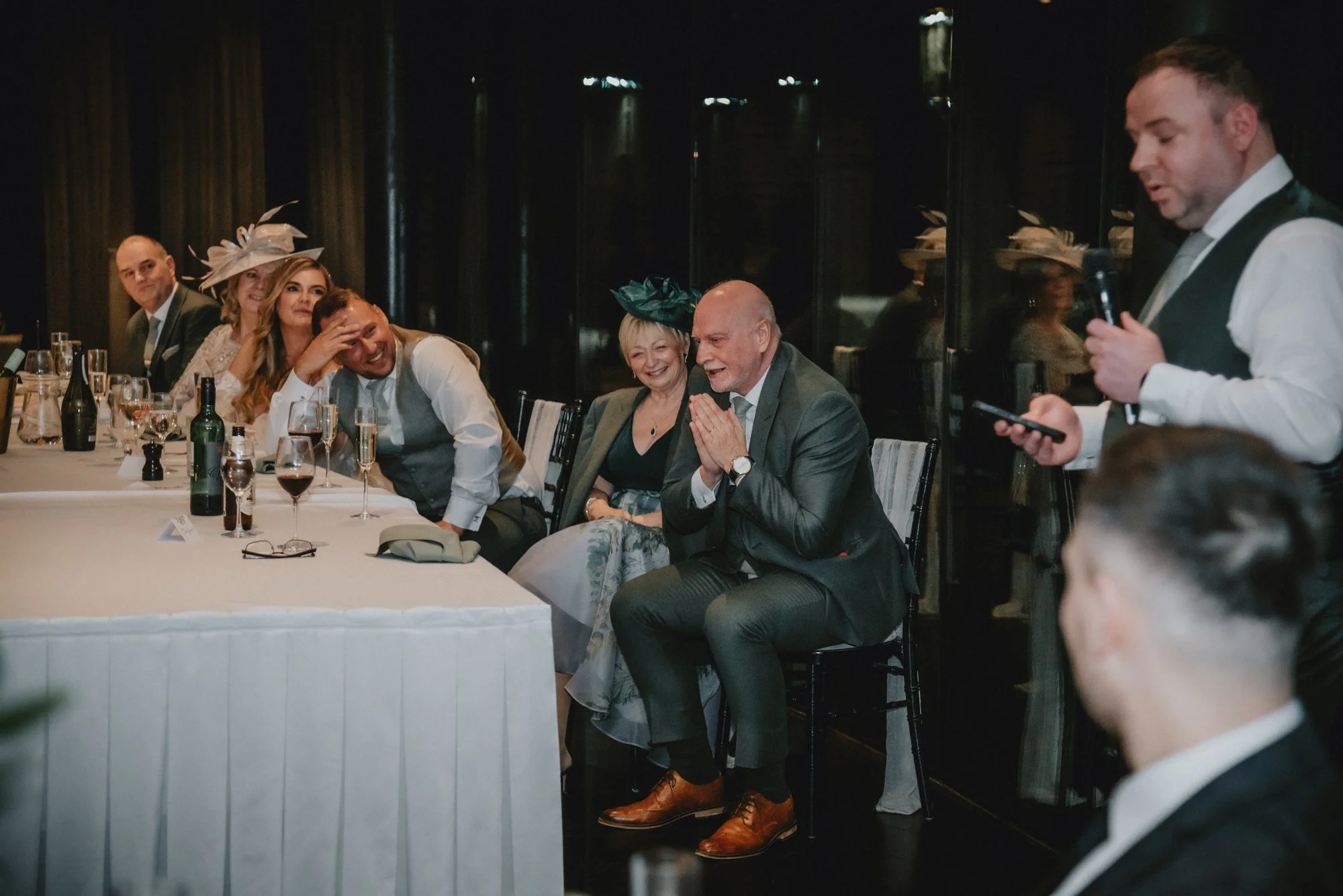 Group of people sitting at a dining table, watching a man giving a speech at a formal event or celebration. The group includes men and women, some wearing hats, with glasses and bottles of wine on the table.