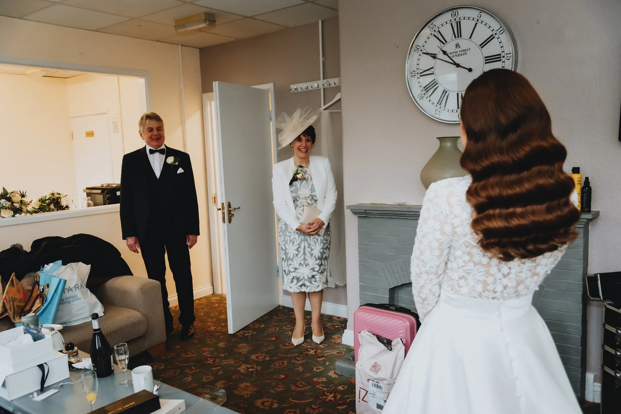 A bride with wavy brown hair in a lace white dress stands in front of two guests, a man in a black tuxedo and a woman in a floral dress with a large hat, in a room with a large clock on the wall.