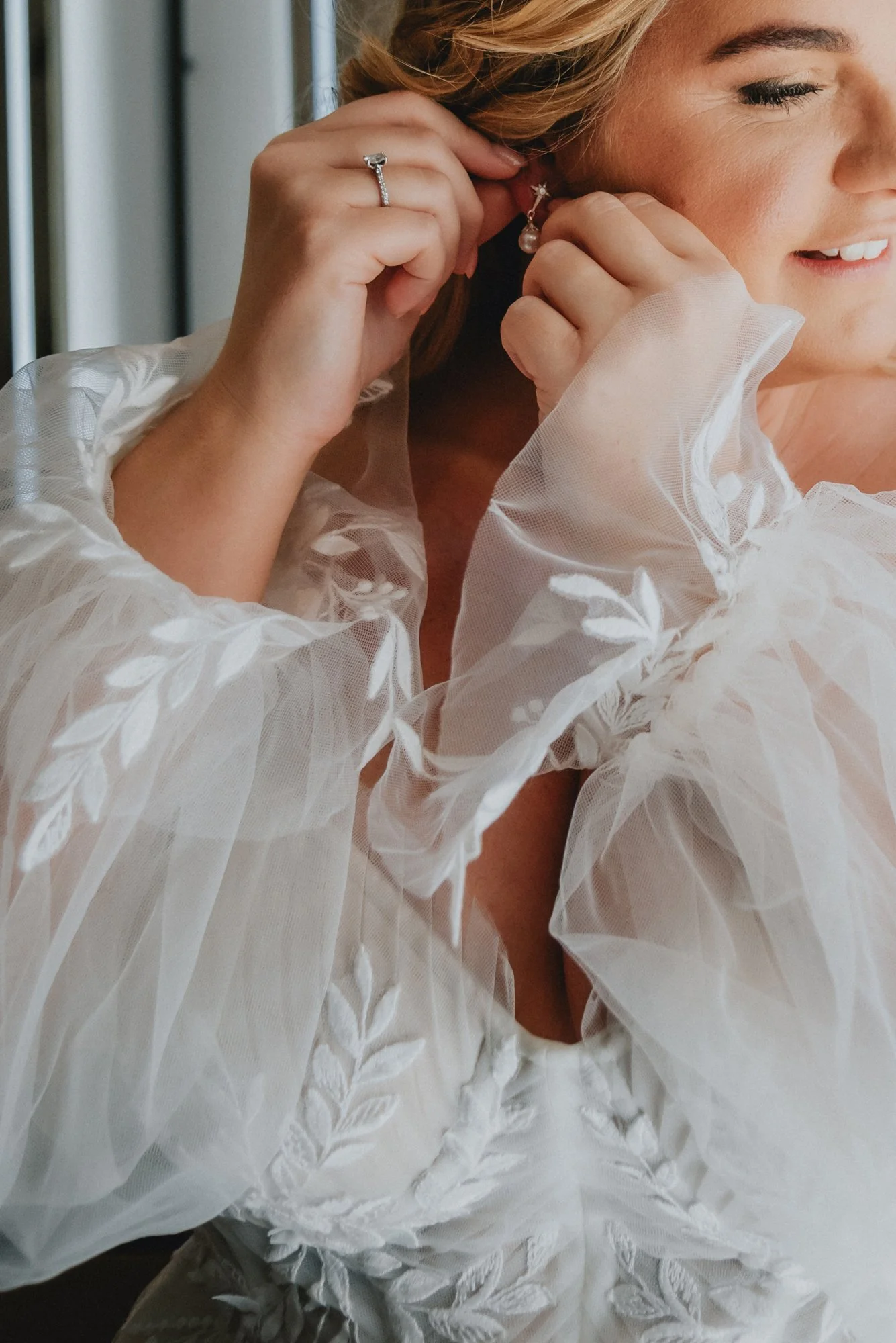 A woman is putting on an earring, wearing a wedding dress with sheer, embroidered sleeves. She has a ring on her finger and is smiling.