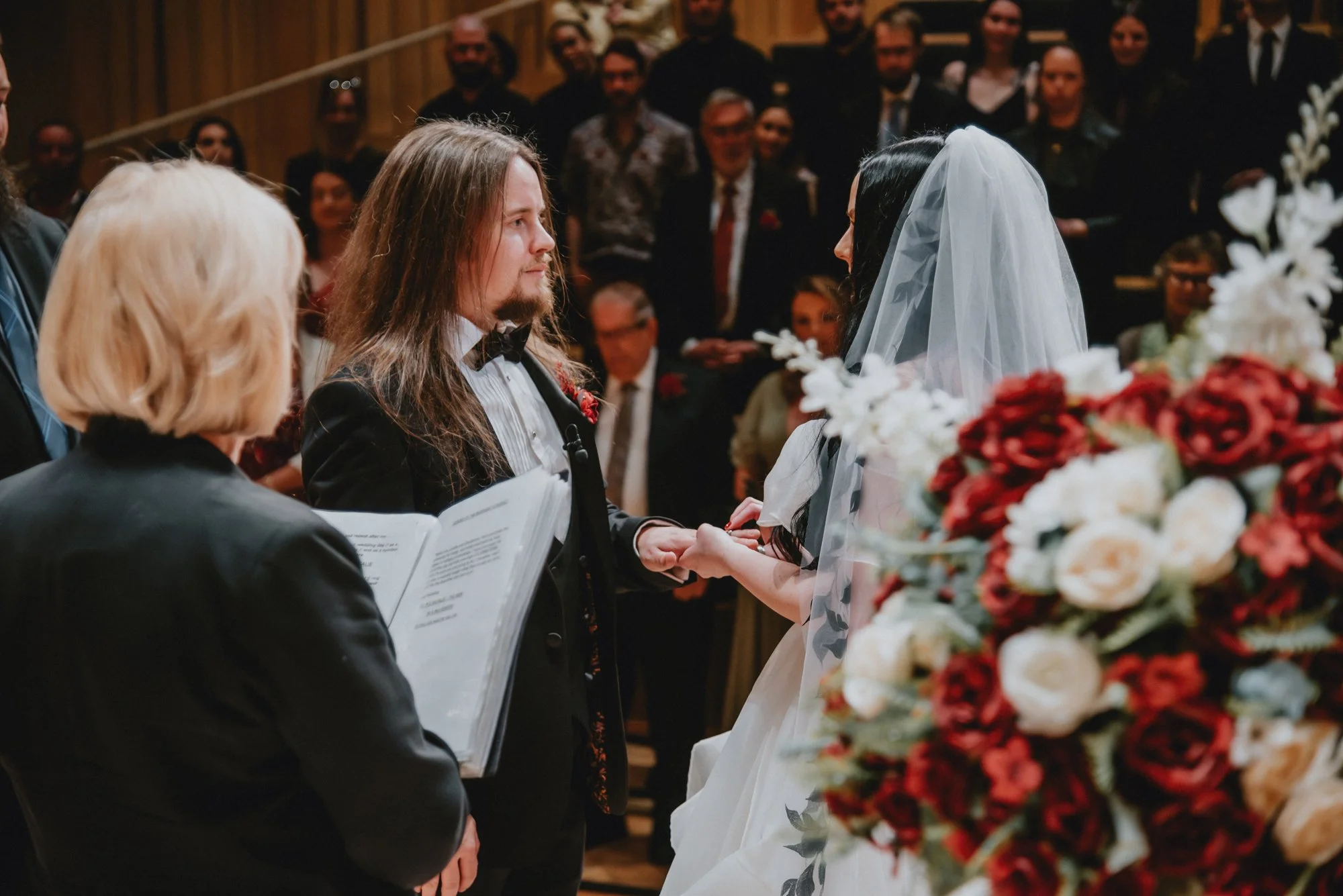 A wedding ceremony with a bride and groom exchanging vows, holding hands, and facing each other. The bride is wearing a white veil, and the groom has long hair, a beard, and is dressed in a tuxedo. An officiant holding a book stands nearby, and a lar