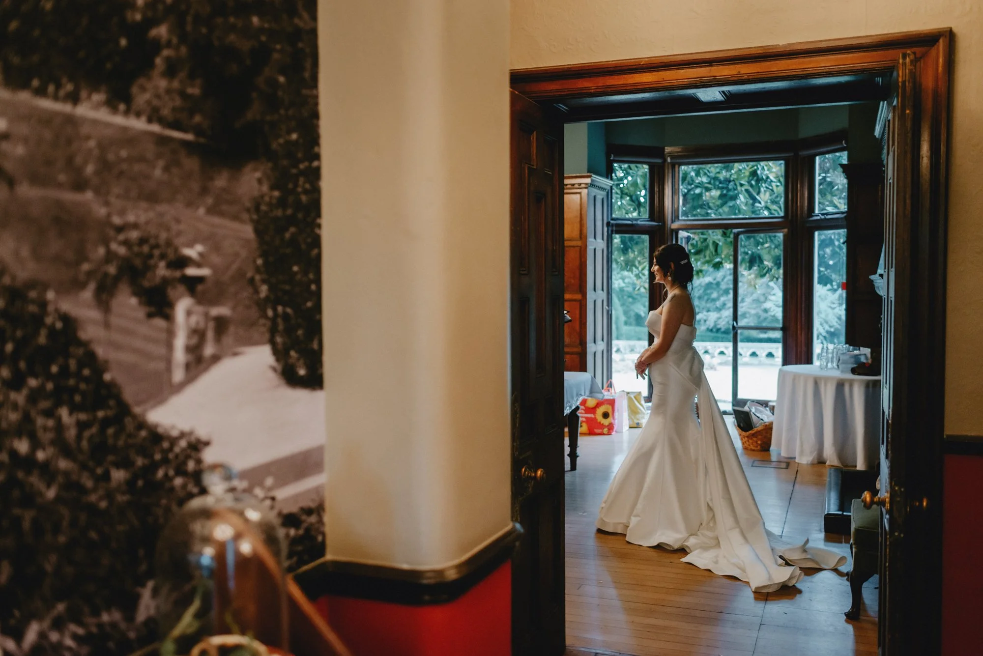 A woman in a white wedding dress standing inside a room, seen through a partially open door, with large windows and greenery outside.