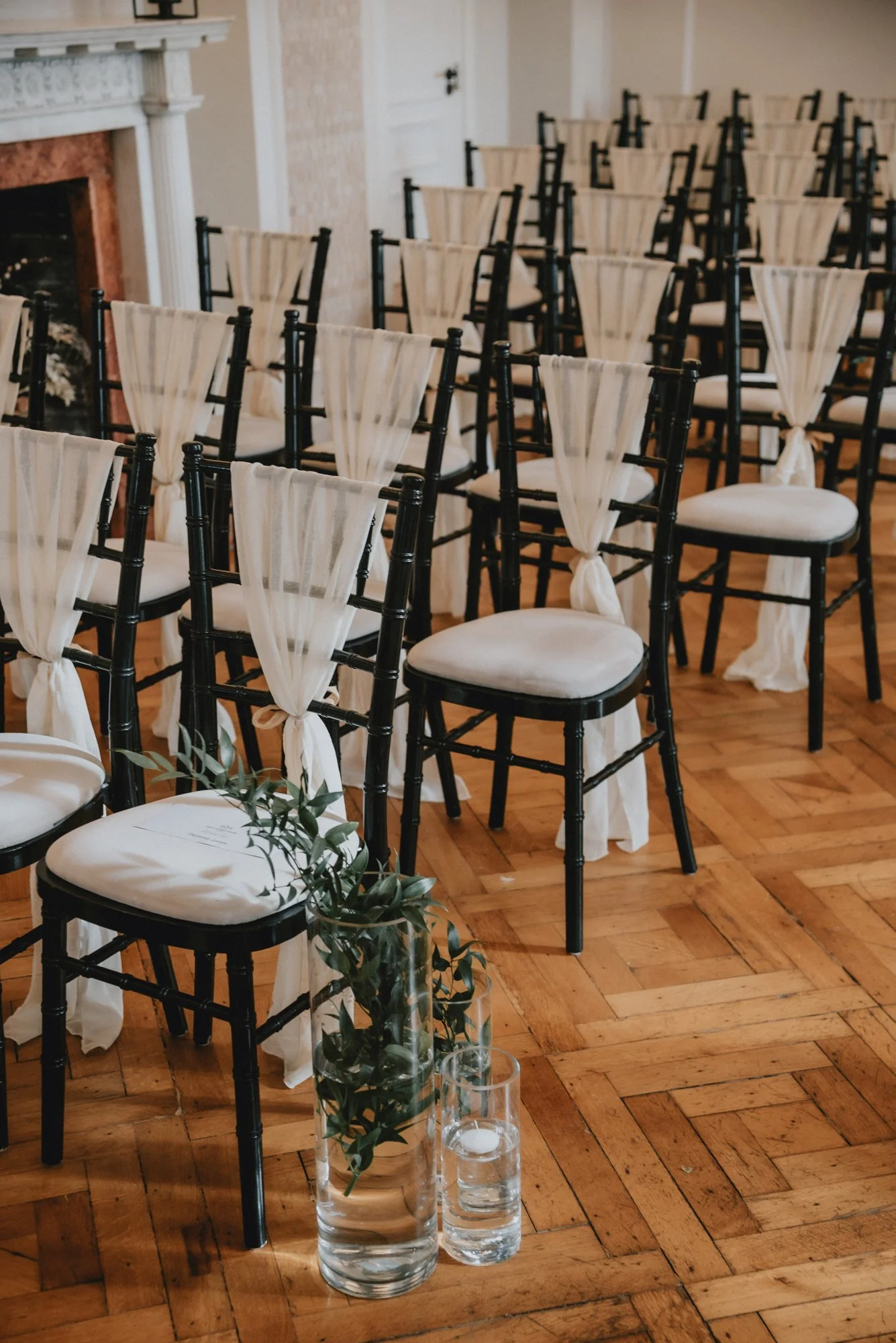 Rows of black chairs with white cushions and white drapes tied to the back in a decorated indoor setting, with a vase of greenery and candles on the wooden floor in the foreground.