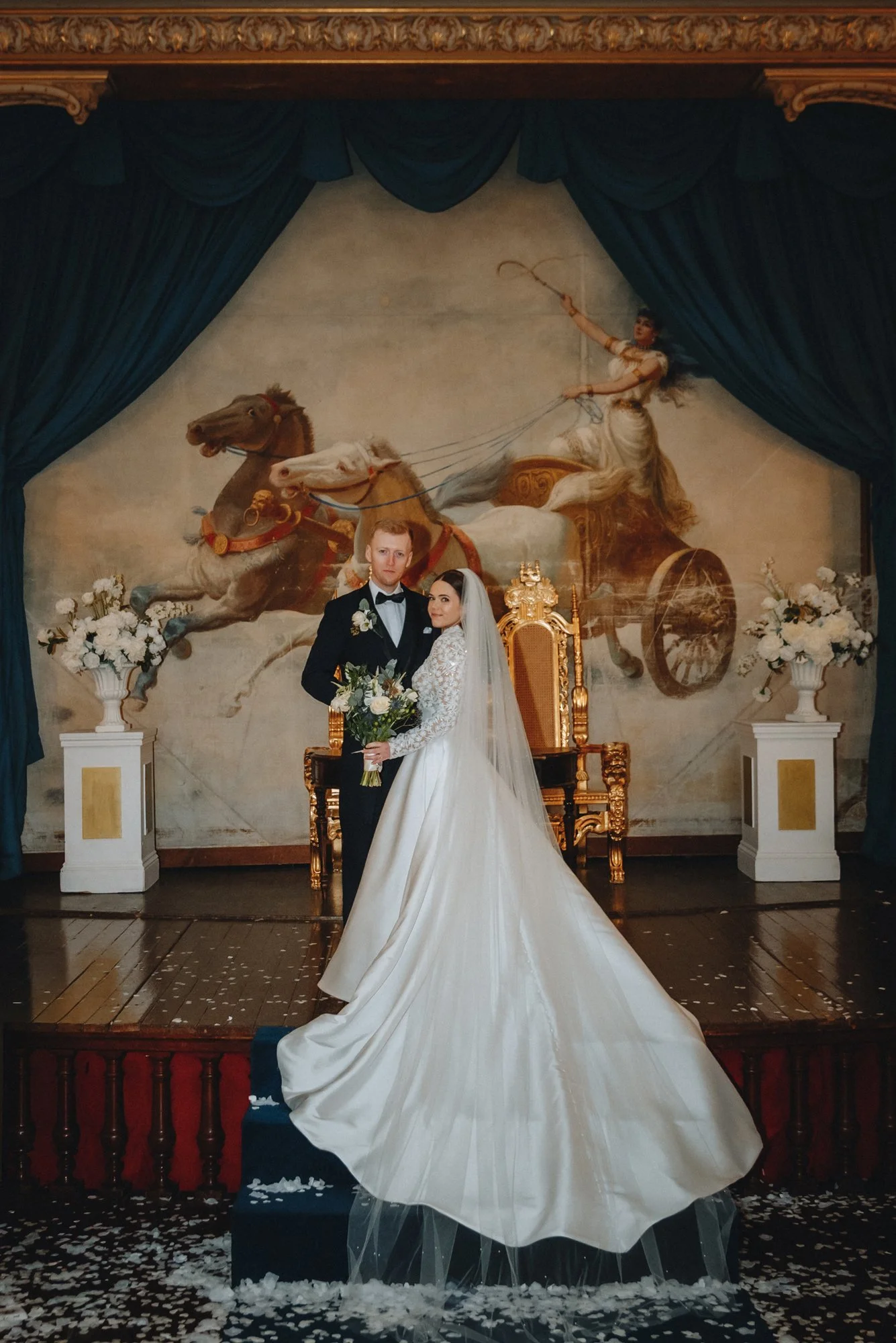 A bride and groom pose on a wedding stage with a large classical painting of a chariot drawn by horses in the background. The bride is wearing a long white wedding gown with a train and holding a bouquet, and the groom is in a black tuxedo. The stage