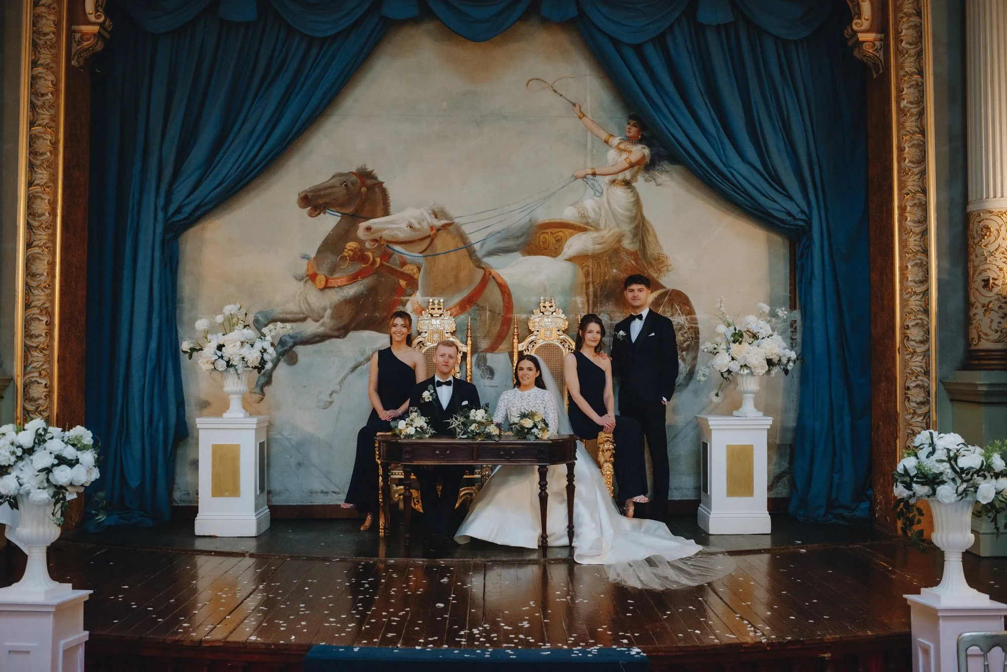 A wedding party posing in front of a classical painting of a woman riding a chariot pulled by horses, with decorated flowers on pedestals and a blue curtain framing the scene.