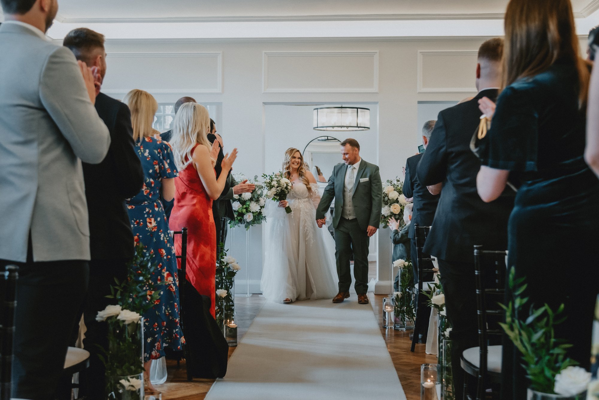 A bride and groom holding hands and walking down the aisle surrounded by guests clapping and celebrating during a wedding ceremony in a bright, elegant room decorated with flowers.