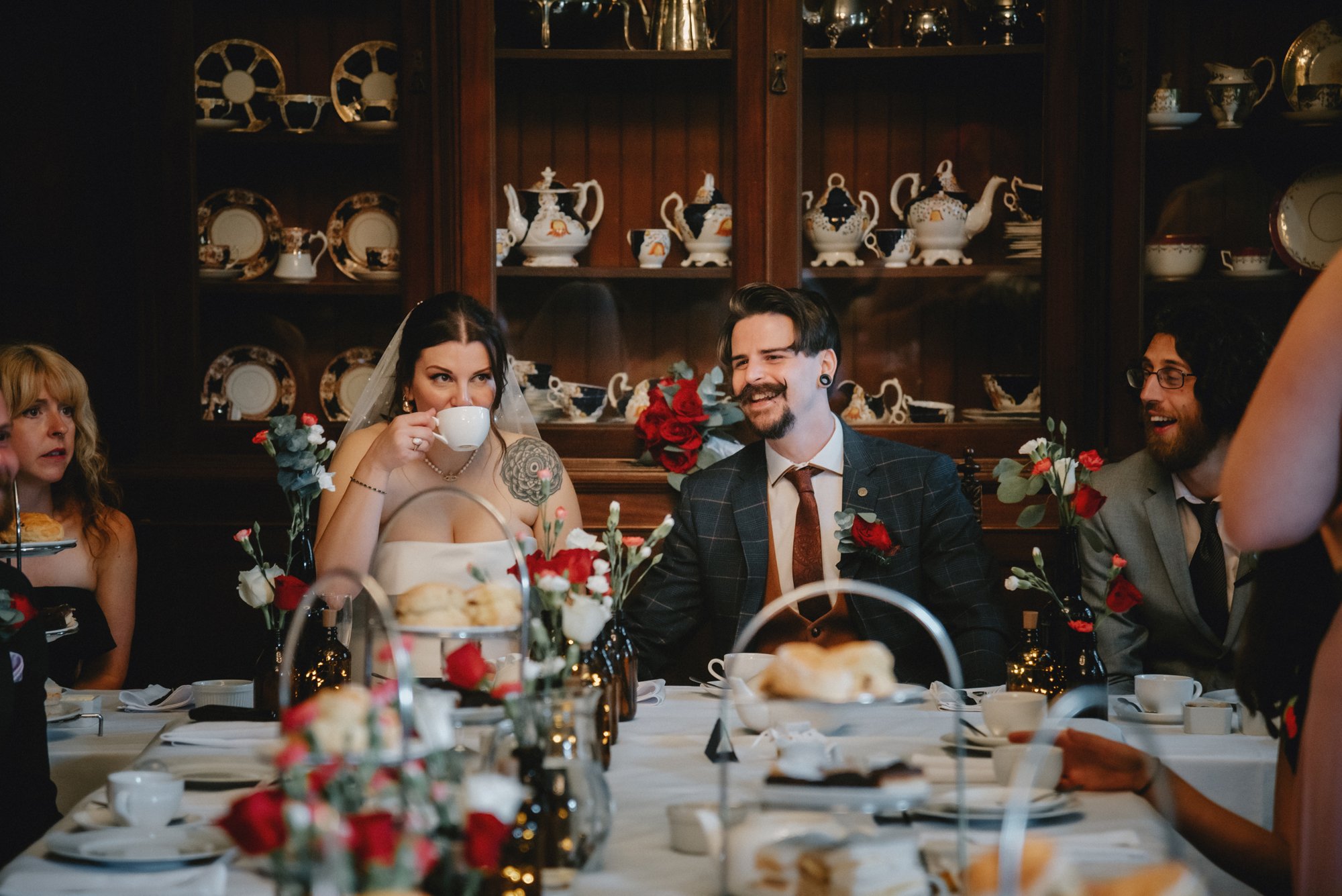People at a wedding reception sitting at a table with flowers, teapots, and cups, with a woman in a wedding dress drinking tea and a man in a suit smiling.