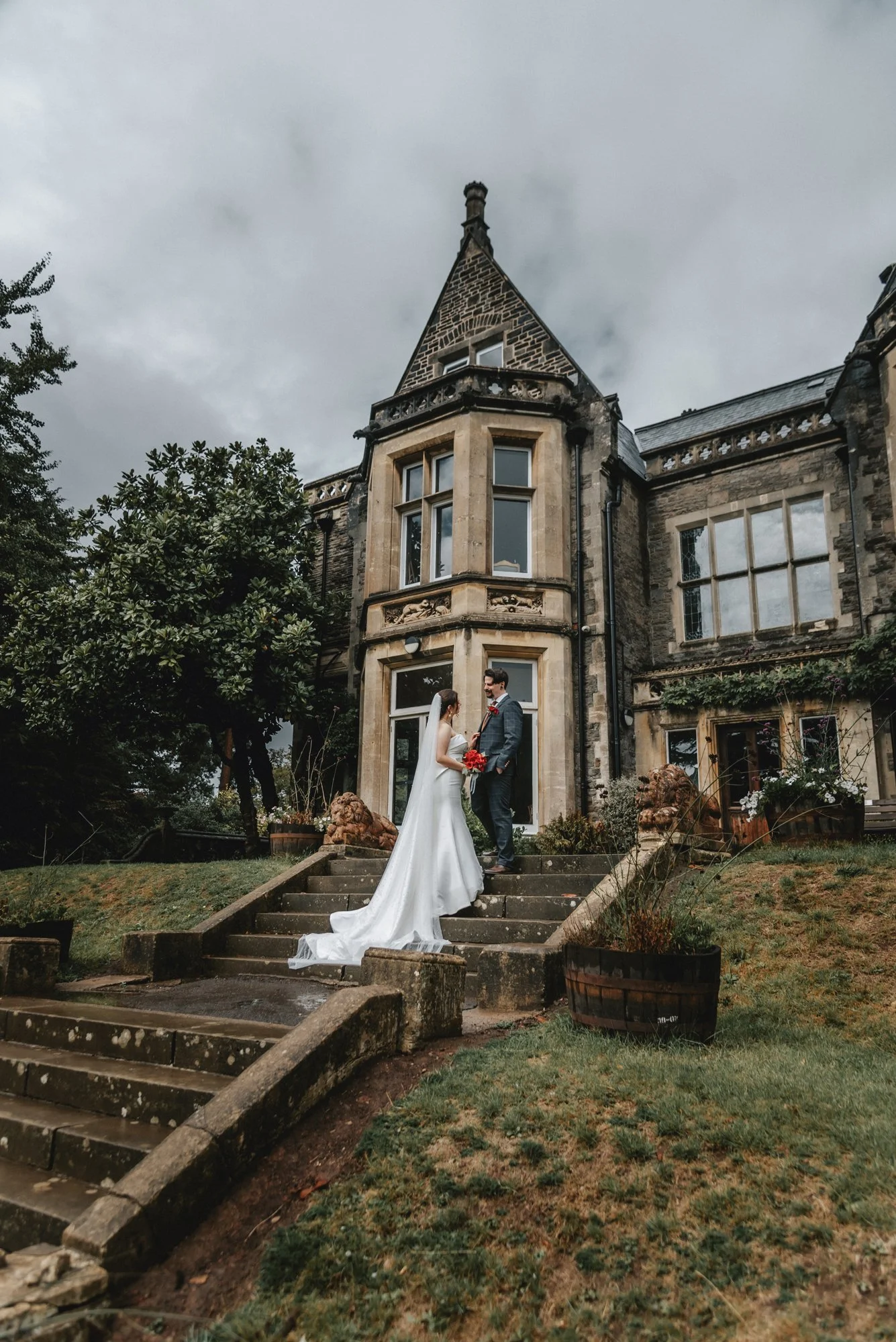 Bride and groom standing on stairs in front of an old, castle-like stone house, holding hands and smiling, wedding scene outdoors on a cloudy day.