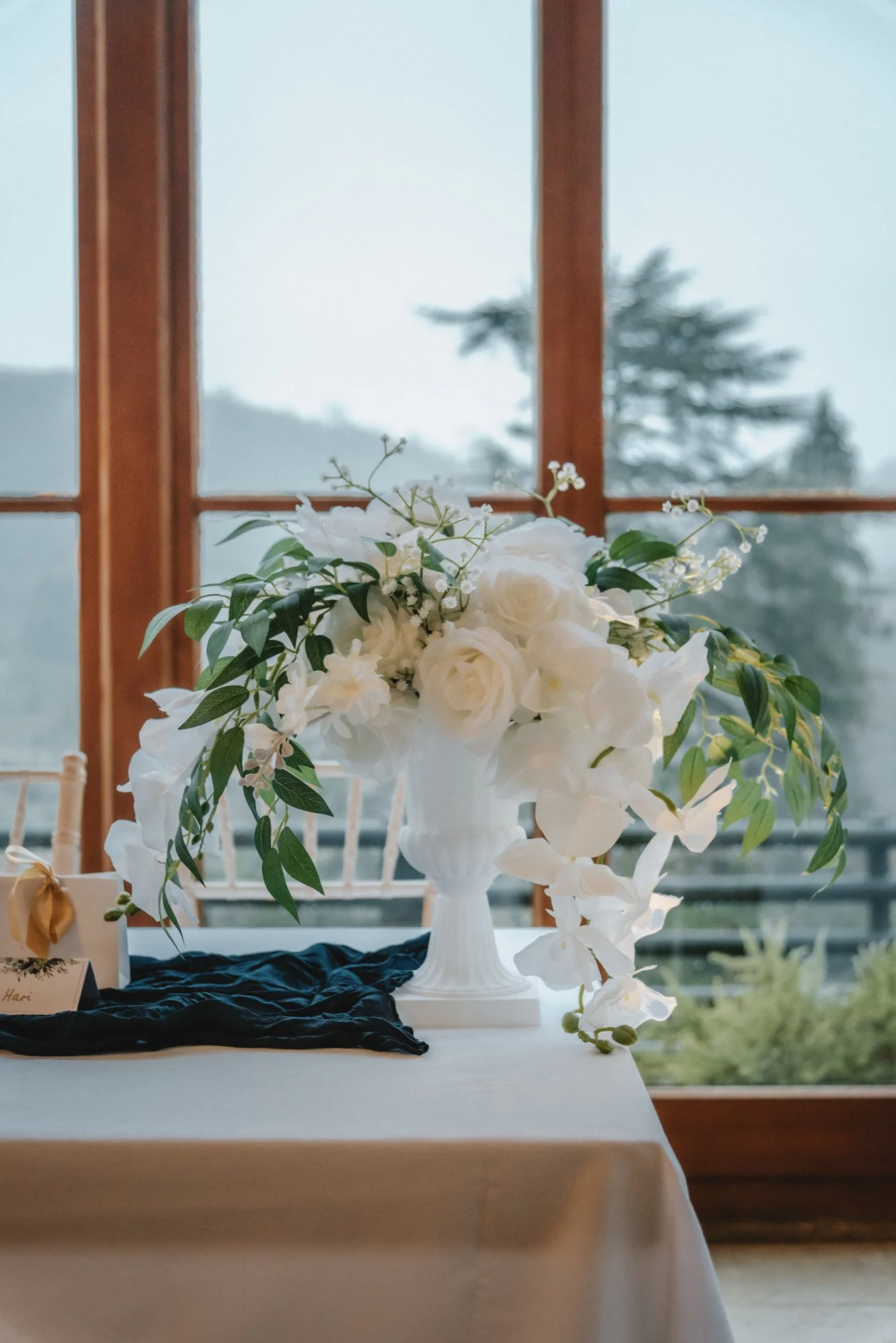 A white floral arrangement with roses and greenery in a white vase on a table, with a foggy outdoor landscape visible through large windows behind it.