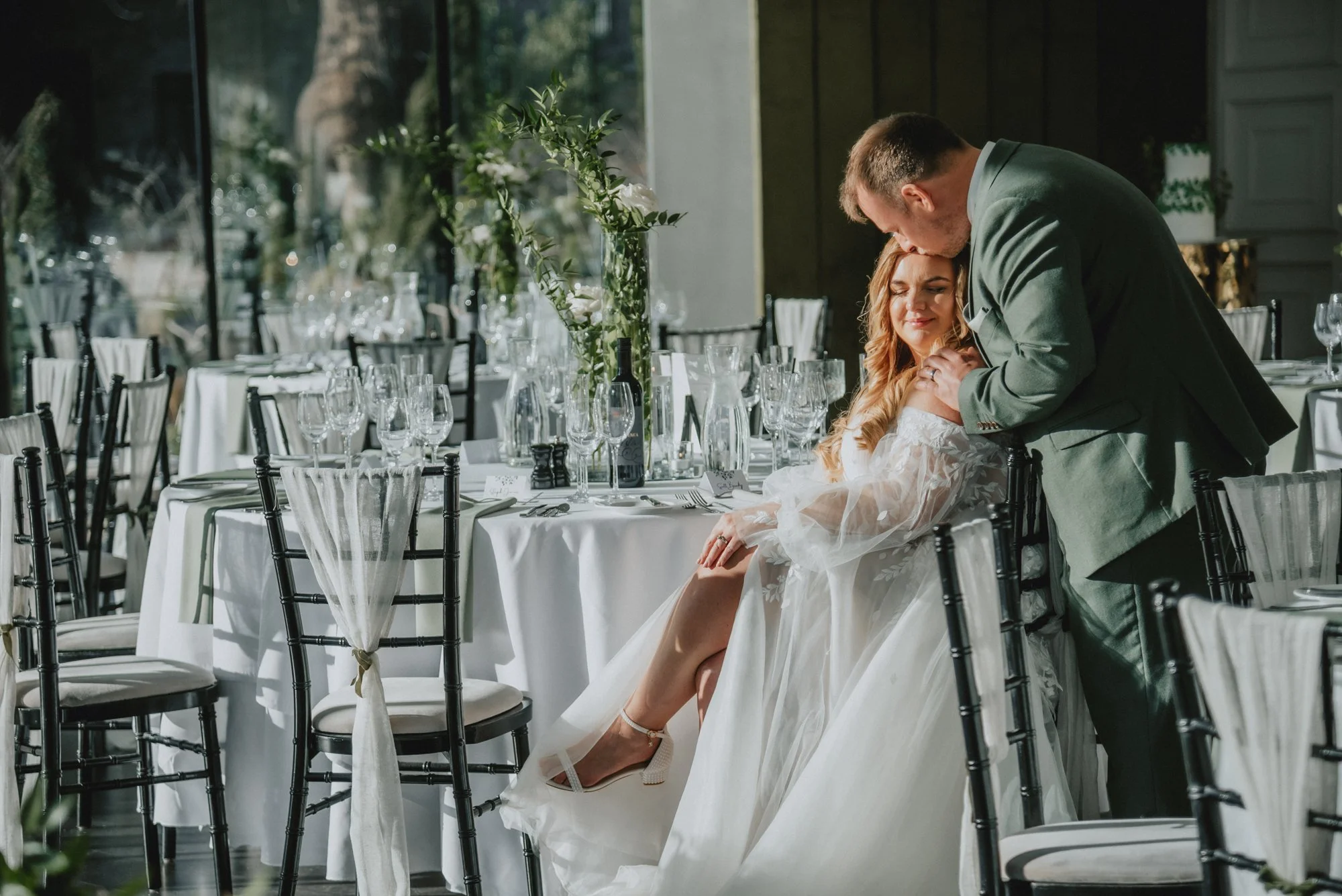 A bride sitting on a chair with her legs crossed, wearing a wedding dress, and a groom leaning over her, kissing her forehead, in a decorated wedding reception hall with tables and chairs.