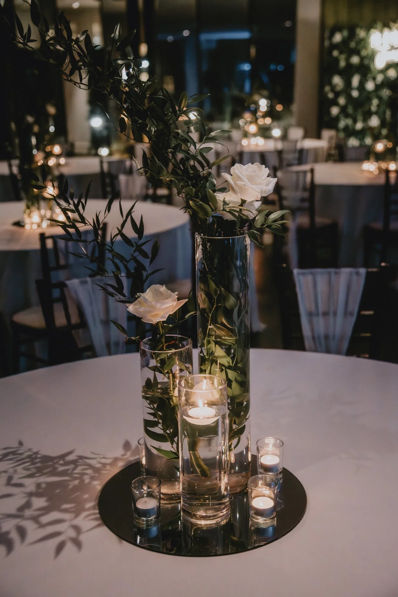 Elegant candle centerpiece with white roses and greenery in glass vases on a round table at night, with dim lighting and other tables with candles in the background.