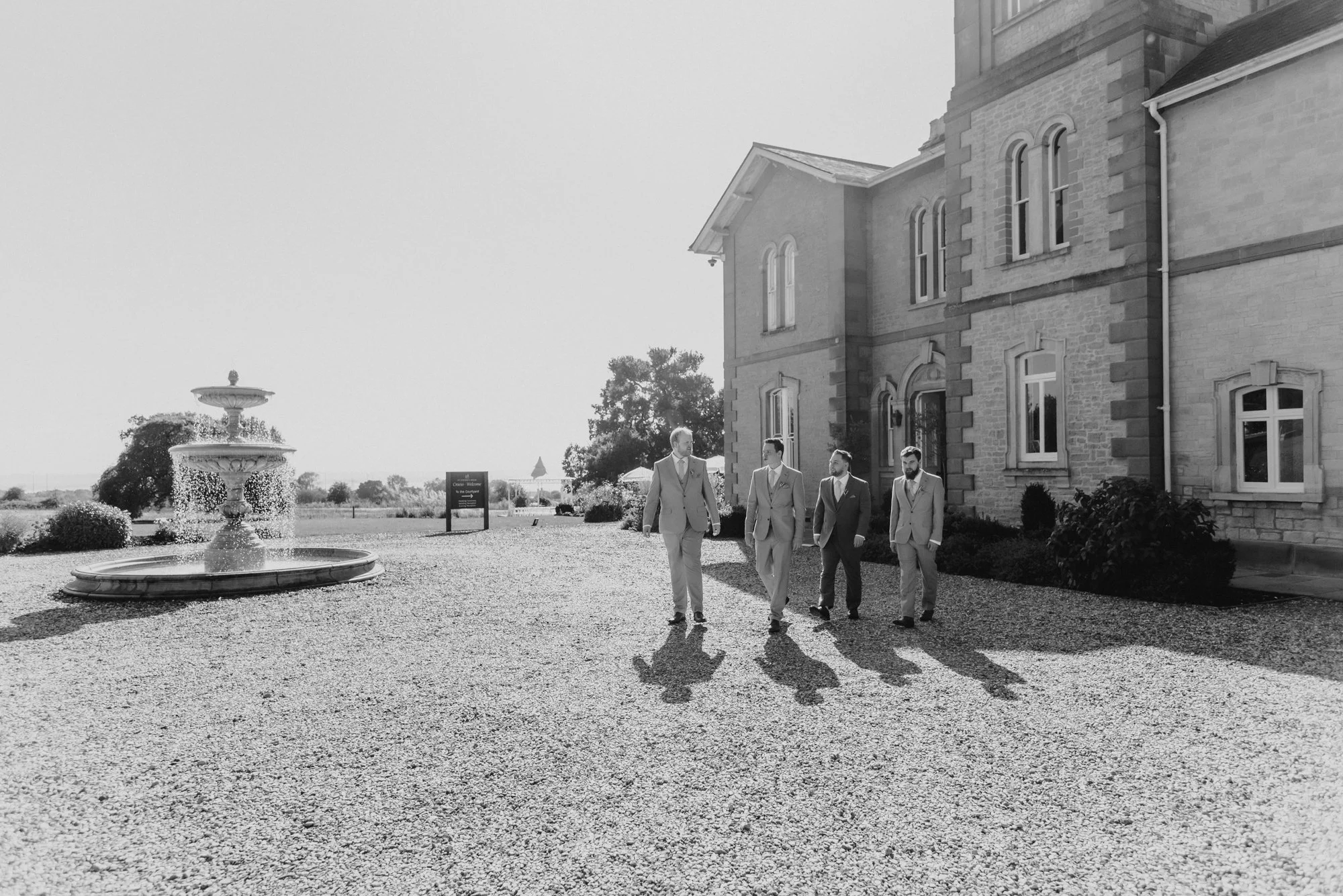 Four men in suits walking and talking outside a large stone building with arched windows and a fountain nearby, sunny day.