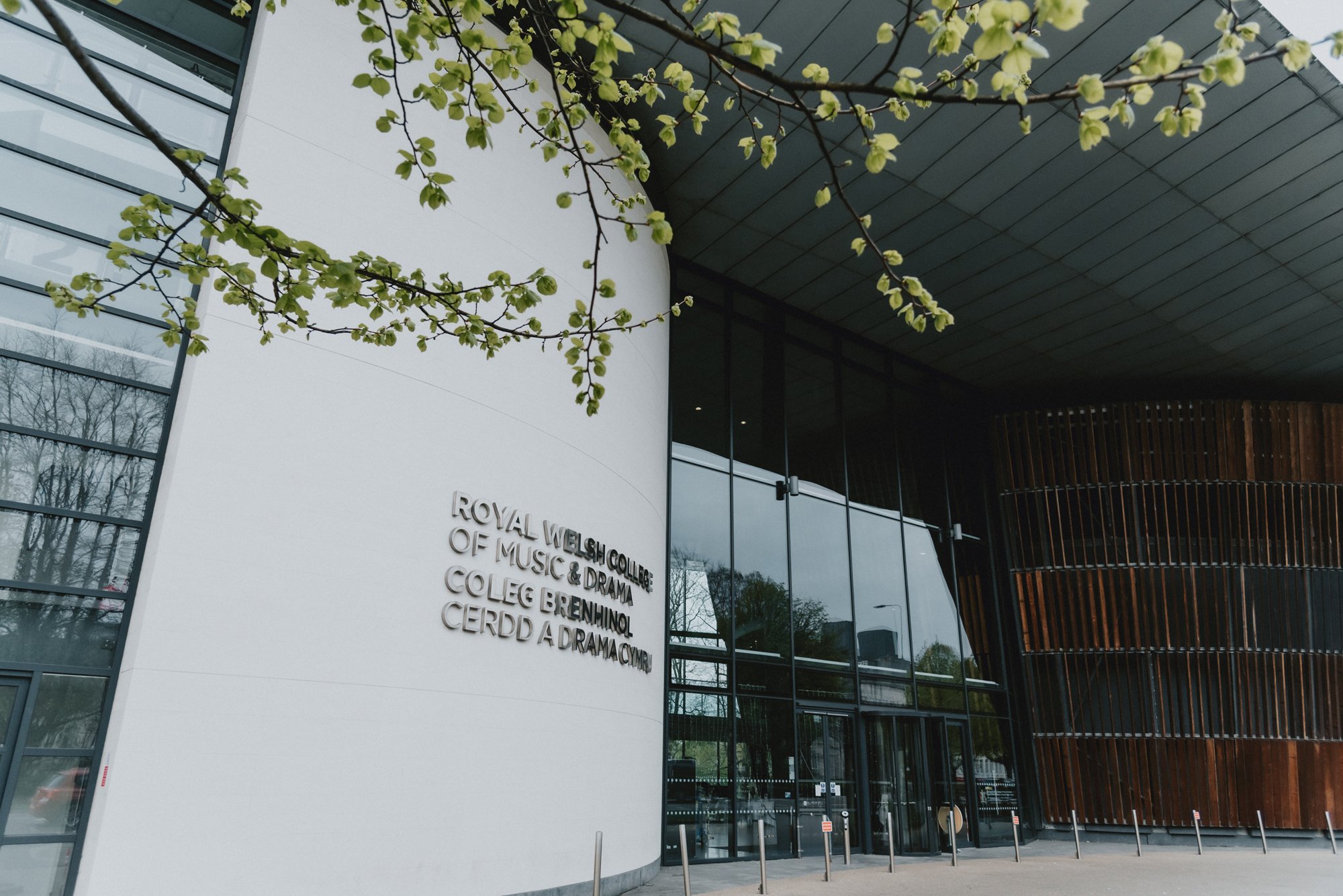 Exterior view of the Royal Welsh College of Music & Drama, showing a modern building with white and wooden paneling, glass doors, and a branch with green leaves in the foreground.