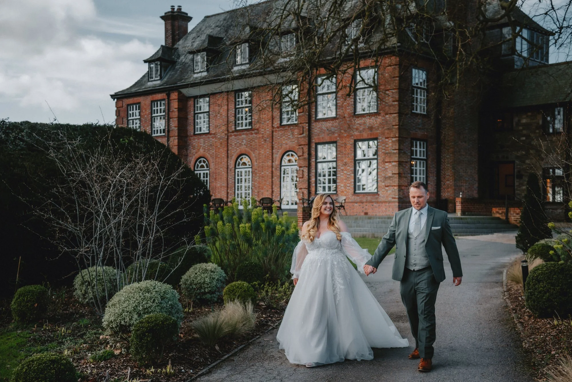 A bride and groom holding hands, walking on a garden path in front of a large brick house, with the bride in a white wedding dress and the groom in a gray suit.
