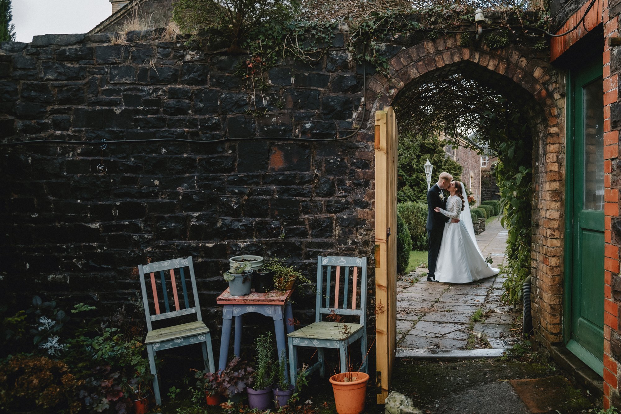 A bride and groom sharing a kiss under an archway in a garden, with a pathway and house in the background.