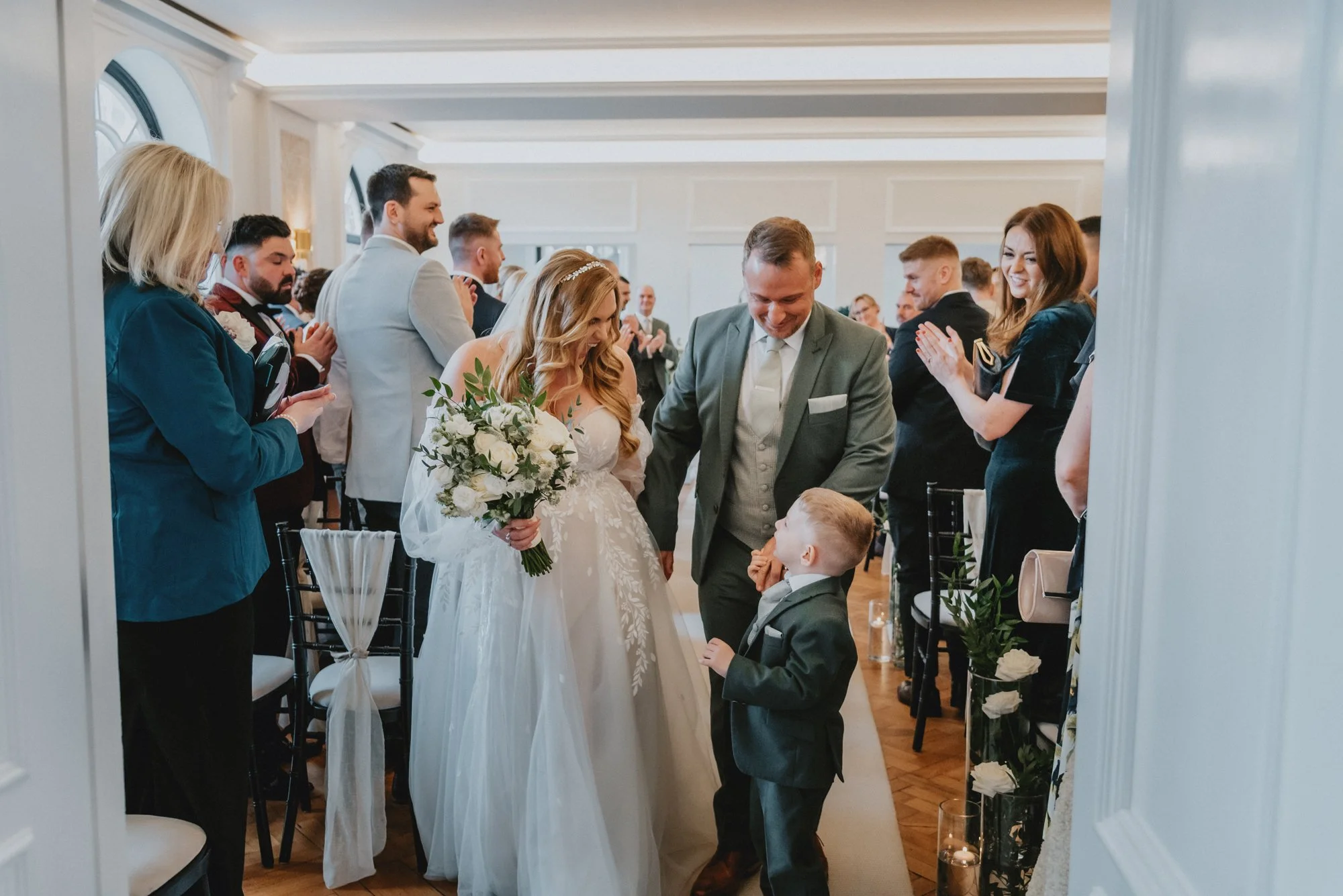 A bride and groom at their wedding ceremony, surrounded by family and friends clapping and celebrating in a decorated indoor venue.