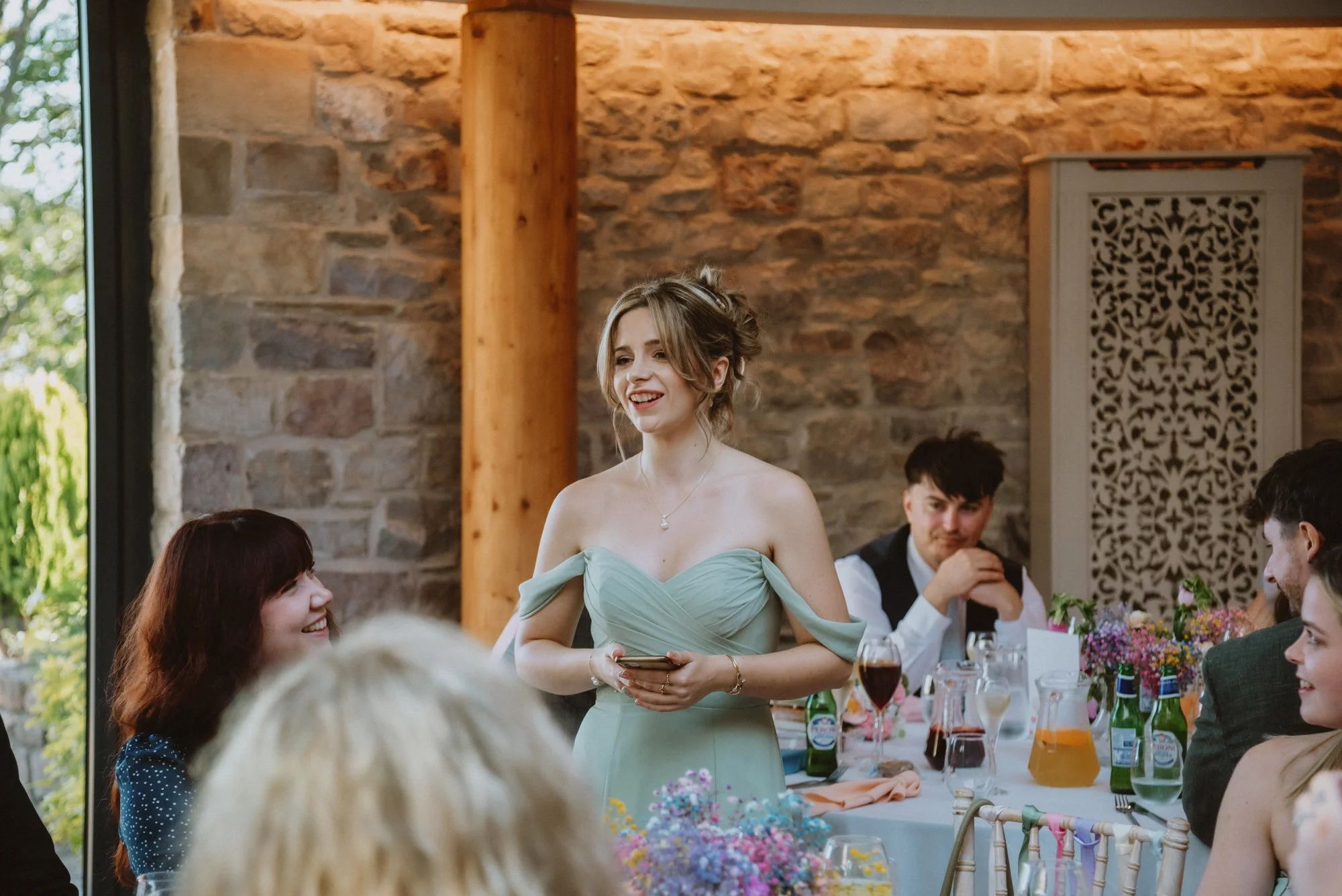 A young woman in a pale green off-shoulder dress speaking at a wedding reception, with guests seated around a decorated table.