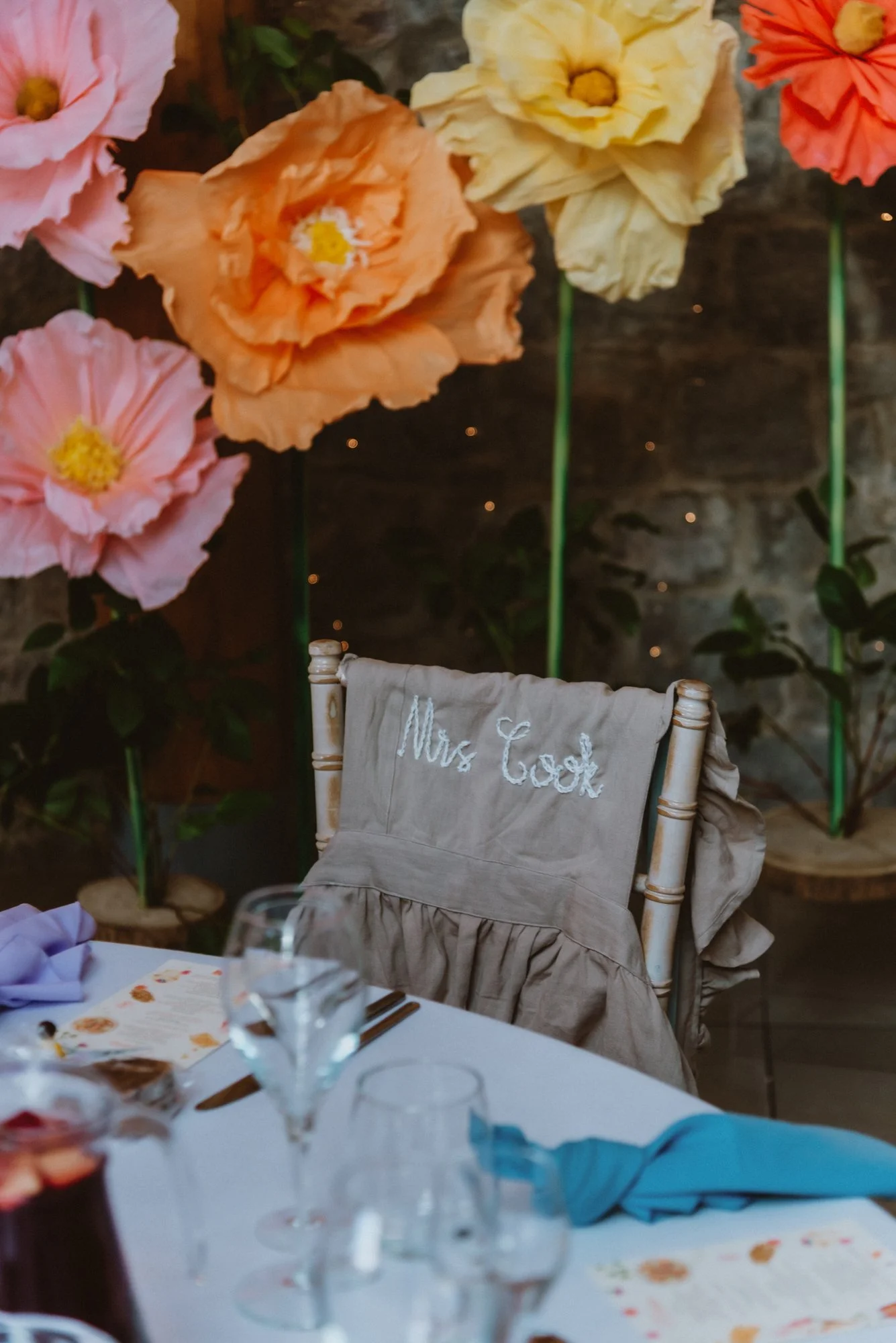 A decorated event table with a chair labeled 'Mrs. Cook' in the foreground and large paper flowers in pink, orange, and yellow in the background.