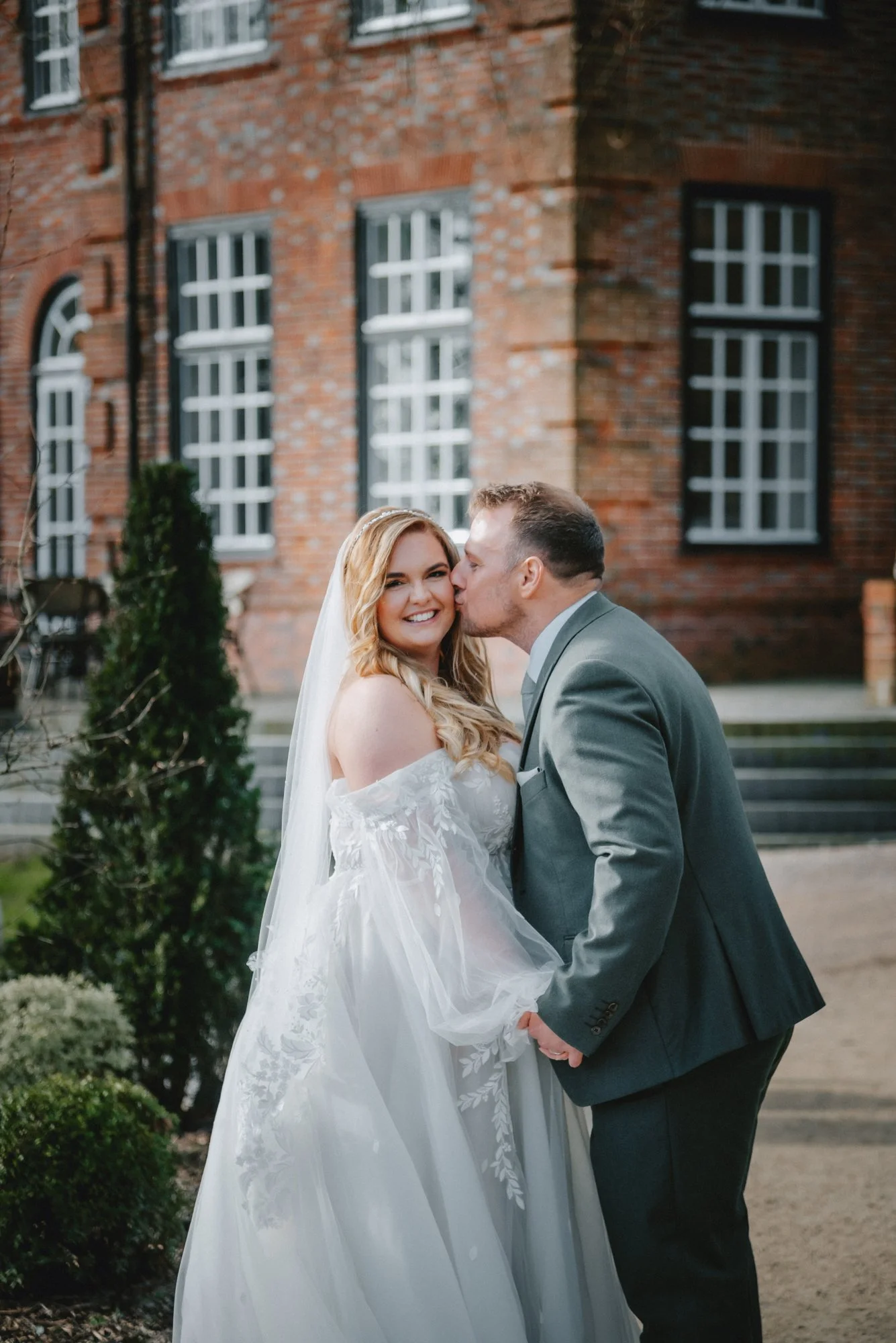 A newlywed couple holding hands, with the groom kissing the bride on the cheek in front of a brick building with large windows.