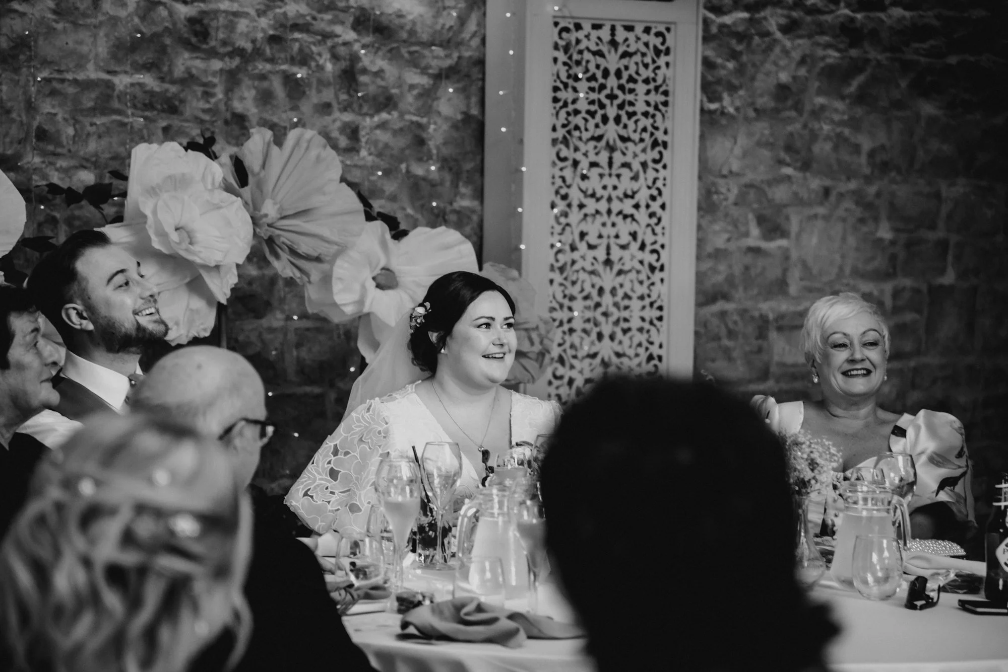 A black and white photo of a group of people sitting at a wedding reception table, smiling and enjoying the event. The background features large decorative flowers and a stone wall.