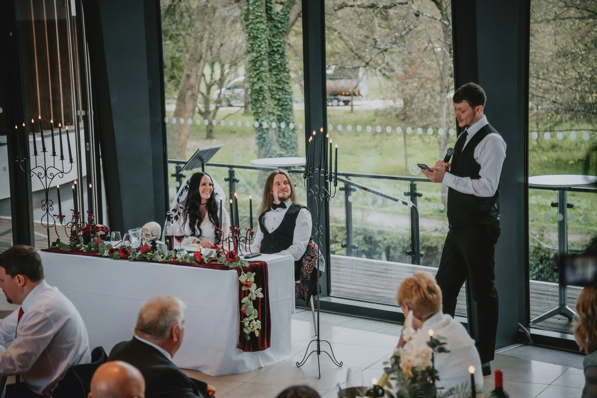 A wedding reception with a bride and groom sitting at a decorated table, a man giving a speech with a microphone, guests seated at tables, and large windows showing an outdoor view of trees and a grassy area.