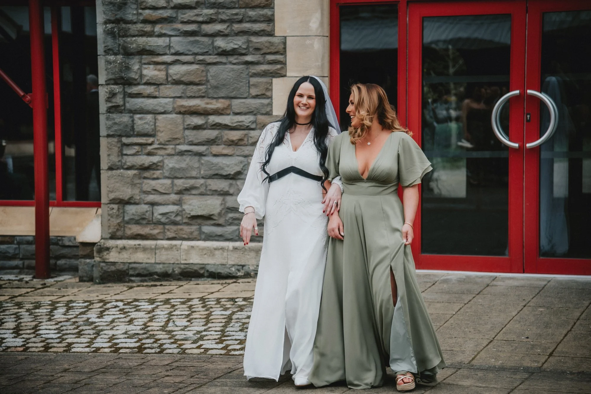 Two women in wedding attire standing on cobblestone sidewalk outside building with brick and stone wall, red doors, smiling and holding hands.