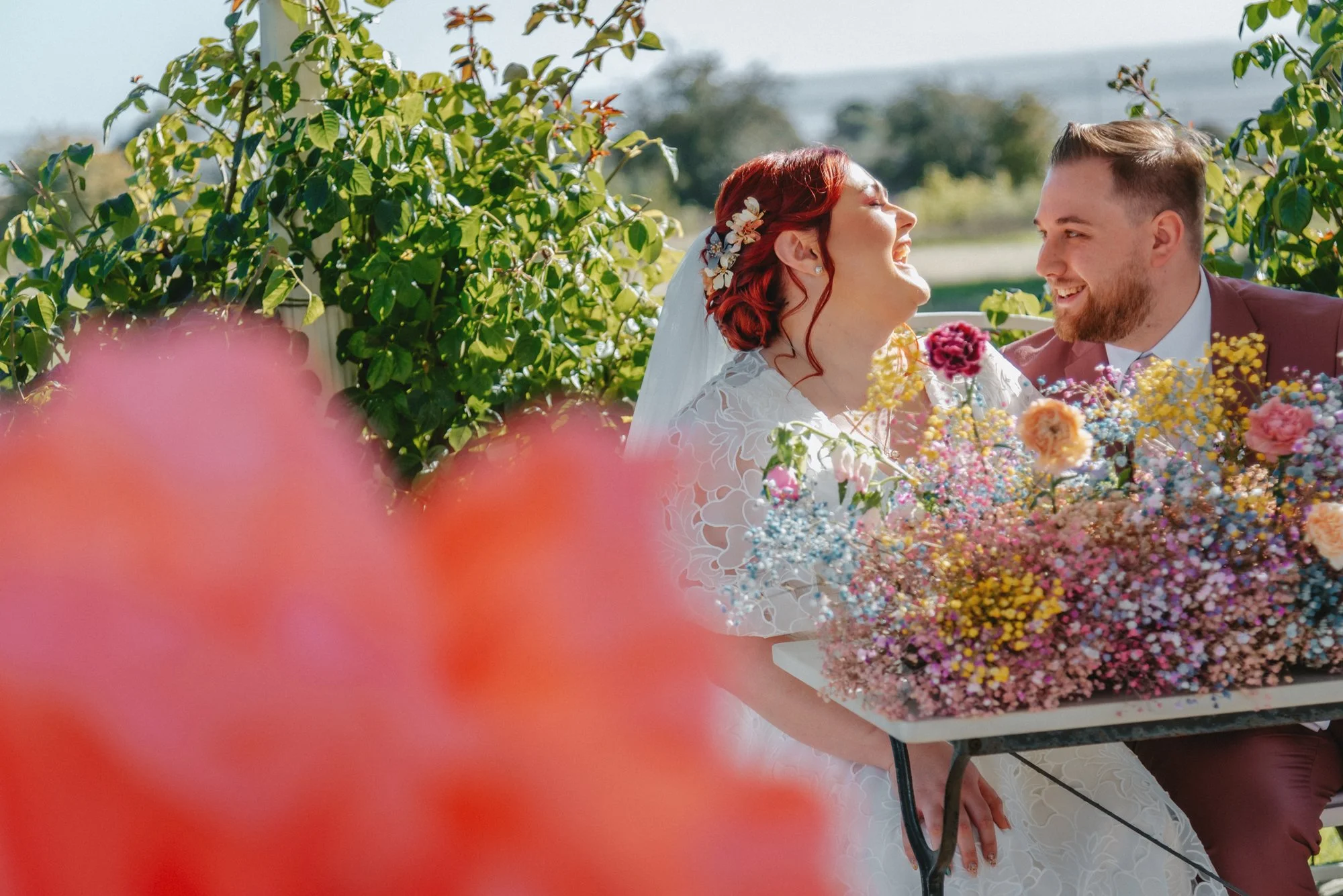 A bride and groom sitting at a table with a large bouquet of colorful flowers, laughing and smiling outdoors with greenery in the background on a sunny day.
