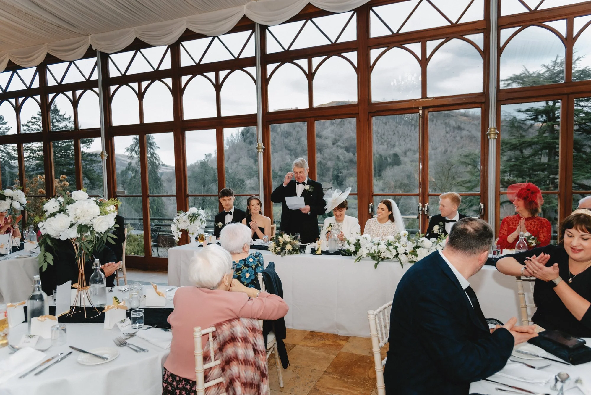 A wedding reception with a woman giving a speech, seated at a decorated head table with the bride and groom, guests clapping, and mountain scenery visible through large windows.