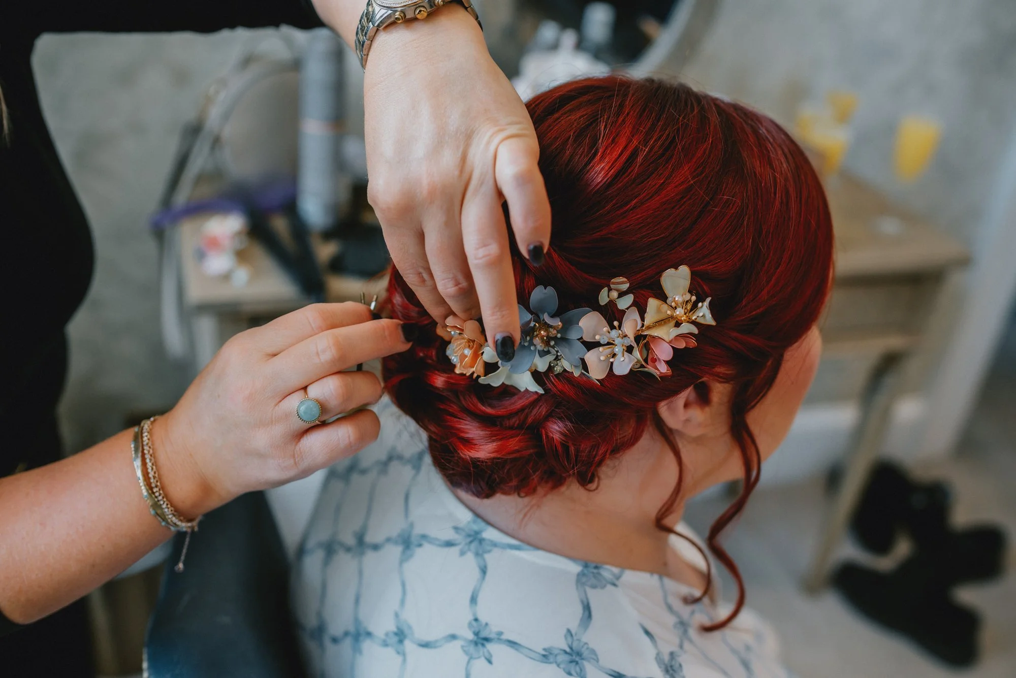 A woman with red hair is getting her hair styled with a floral hair accessory by a stylist. The stylist is adjusting the accessory near the woman's hair.