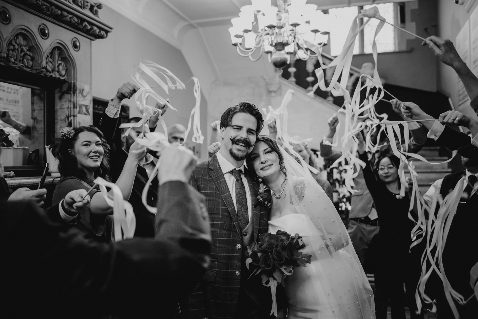 A black and white photo of a newlywed couple smiling and leaning on each other, surrounded by friends and family celebrating with streamers in a decorated indoor venue.