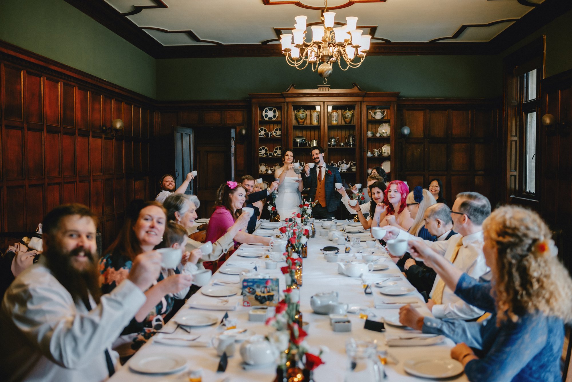 A large group of people gathered around a long table in a wood-paneled dining room, raising bowls in a toast, celebrating a wedding with the bride and groom standing at the head of the table.
