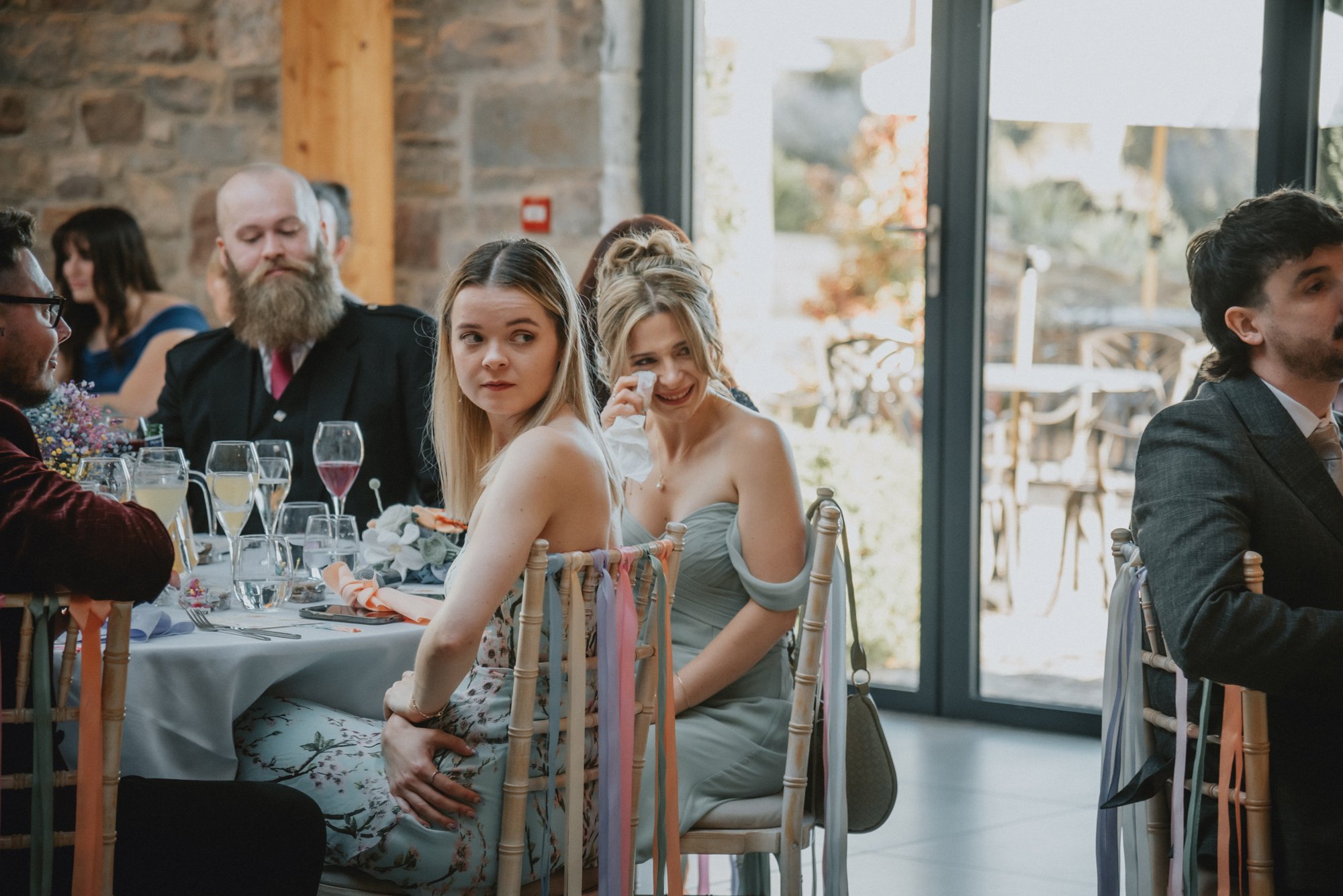 People sitting at a decorated wedding reception table indoors, with glasses and floral arrangements, and large windows showing outdoor scenery.