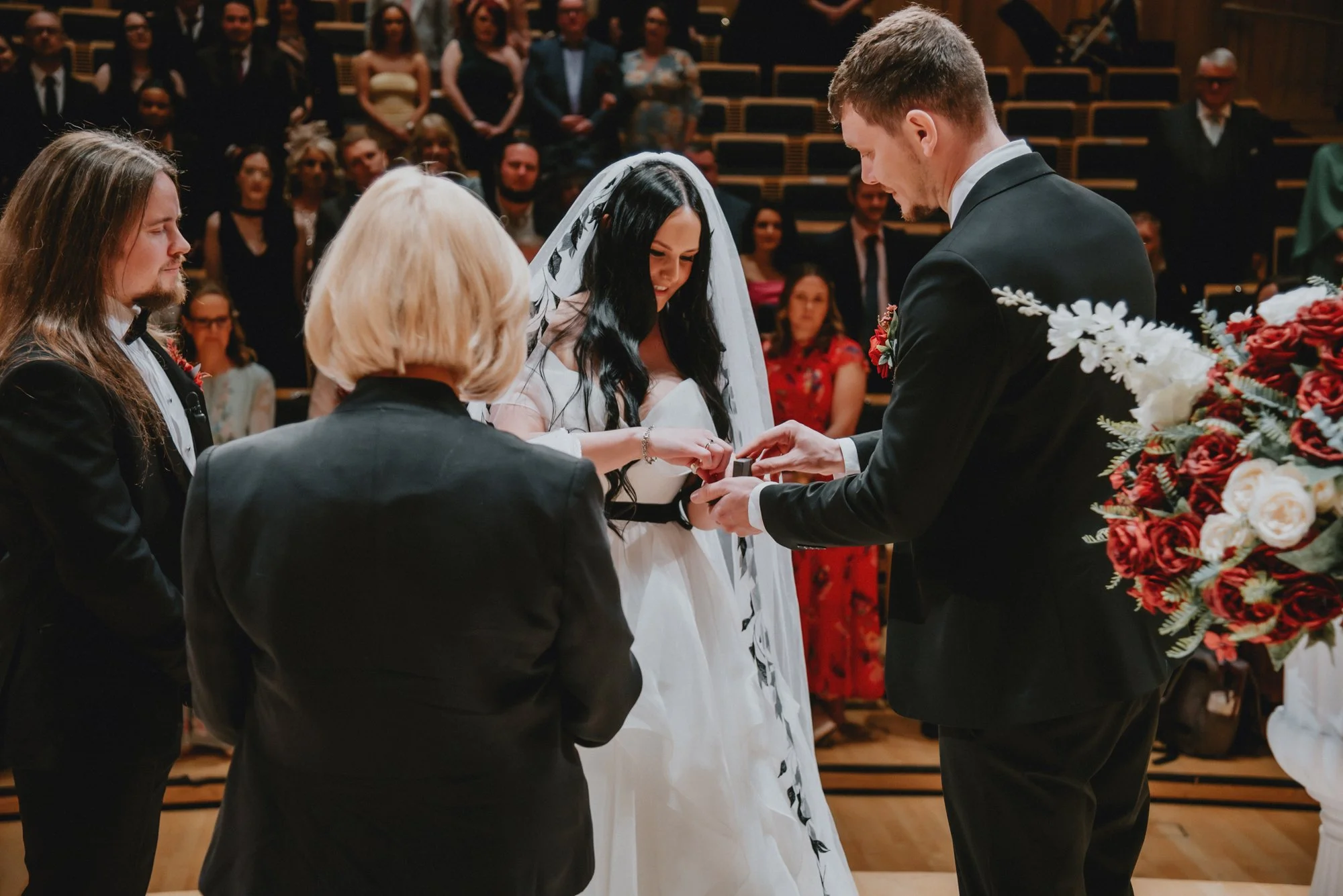 A bride and groom exchange rings during their wedding ceremony, surrounded by attendants and guests in a formal indoor setting with wooden floors and a large floral arrangement.