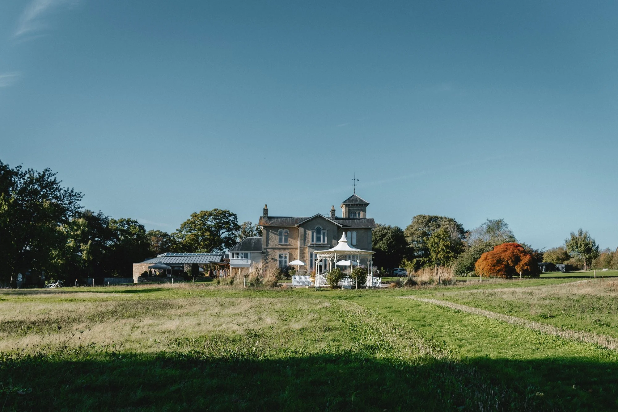A large, historic house surrounded by trees and open grassy fields under a clear blue sky.