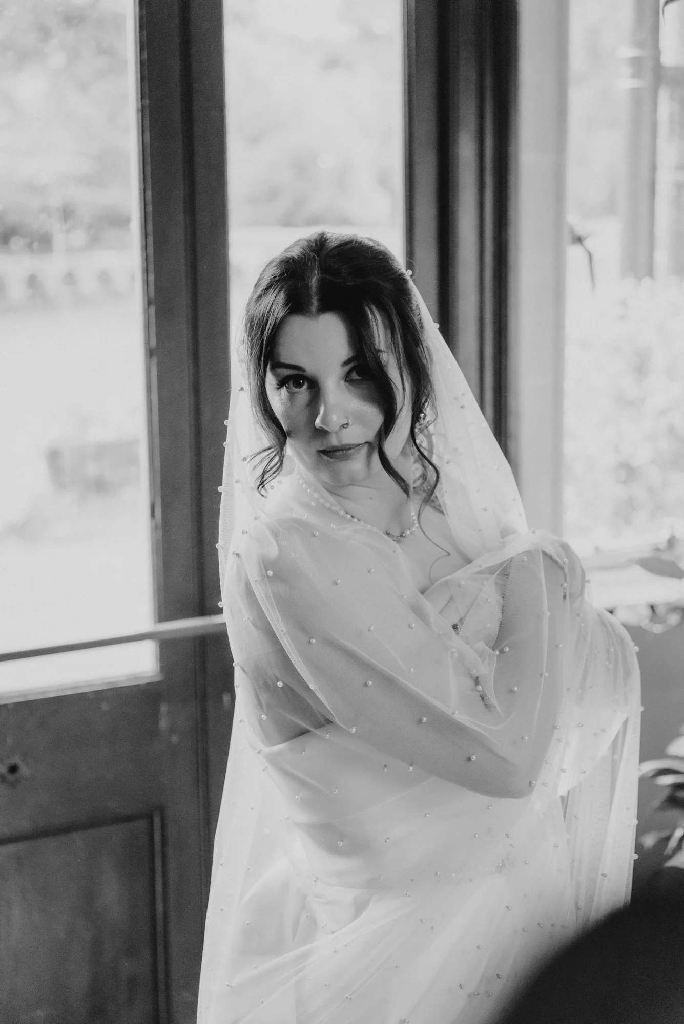 A woman in a wedding dress with a veil standing in front of a window, looking into the camera with a playful expression.