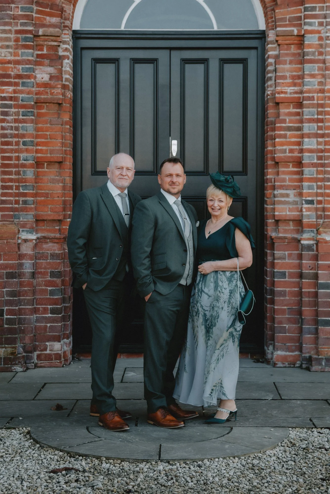 Three people dressed in formal attire standing outside in front of a brick wall and black door.