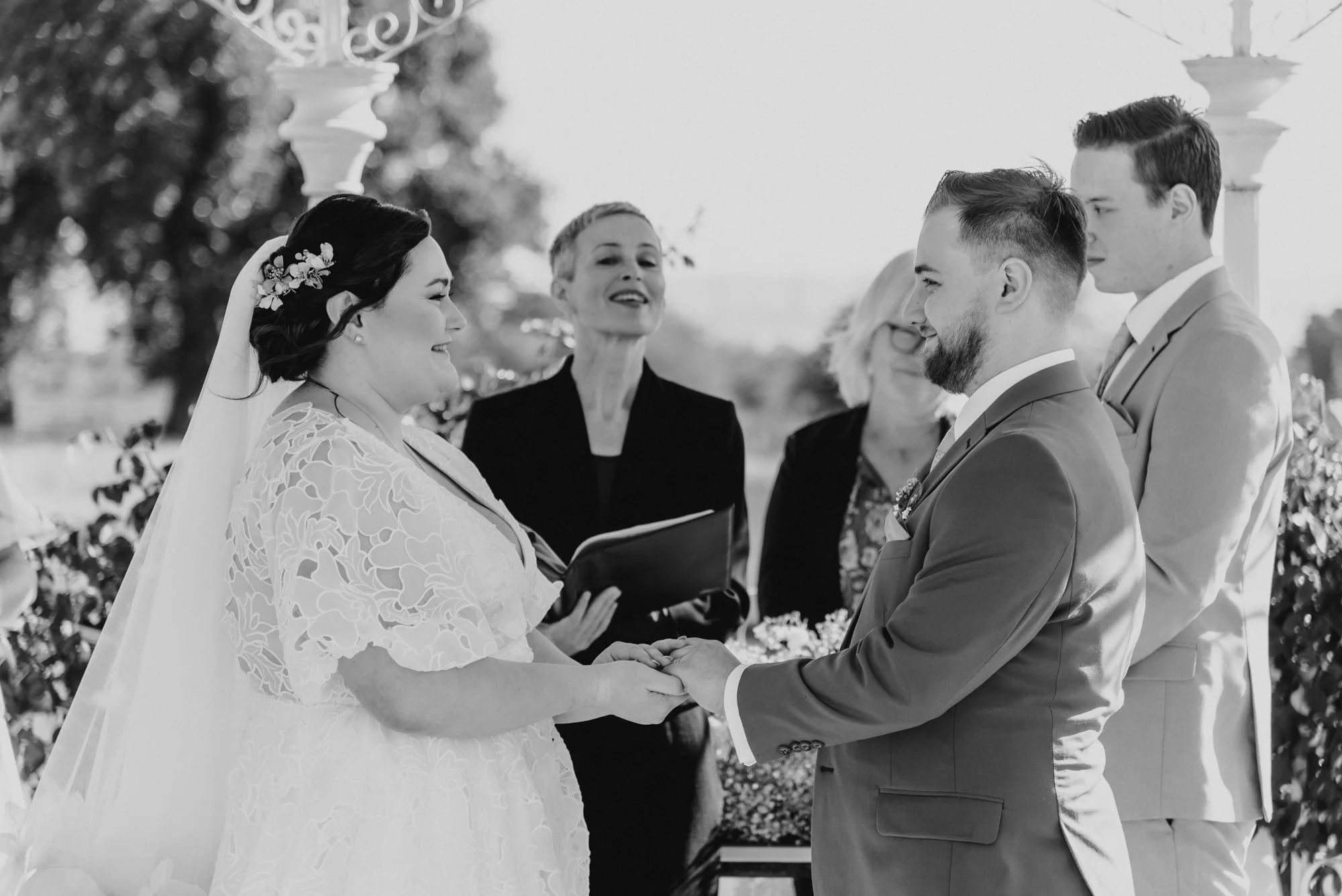 A black and white photo of a wedding ceremony outdoors showing a bride and groom holding hands and smiling at each other, with an officiant and two witnesses or family members standing behind them.