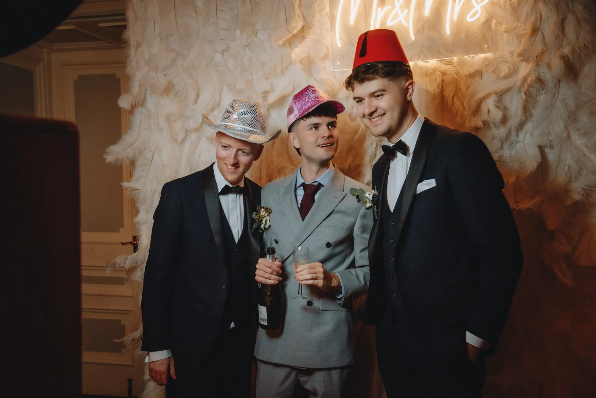 Three young men dressed in tuxedos with boutonnières, wearing humorous hats (a cowboy hat, a pink glittery hat, and a red fez), are at a party or celebration, standing in front of a feathered backdrop with a neon sign that says 'Mr & Mrs.' One of the