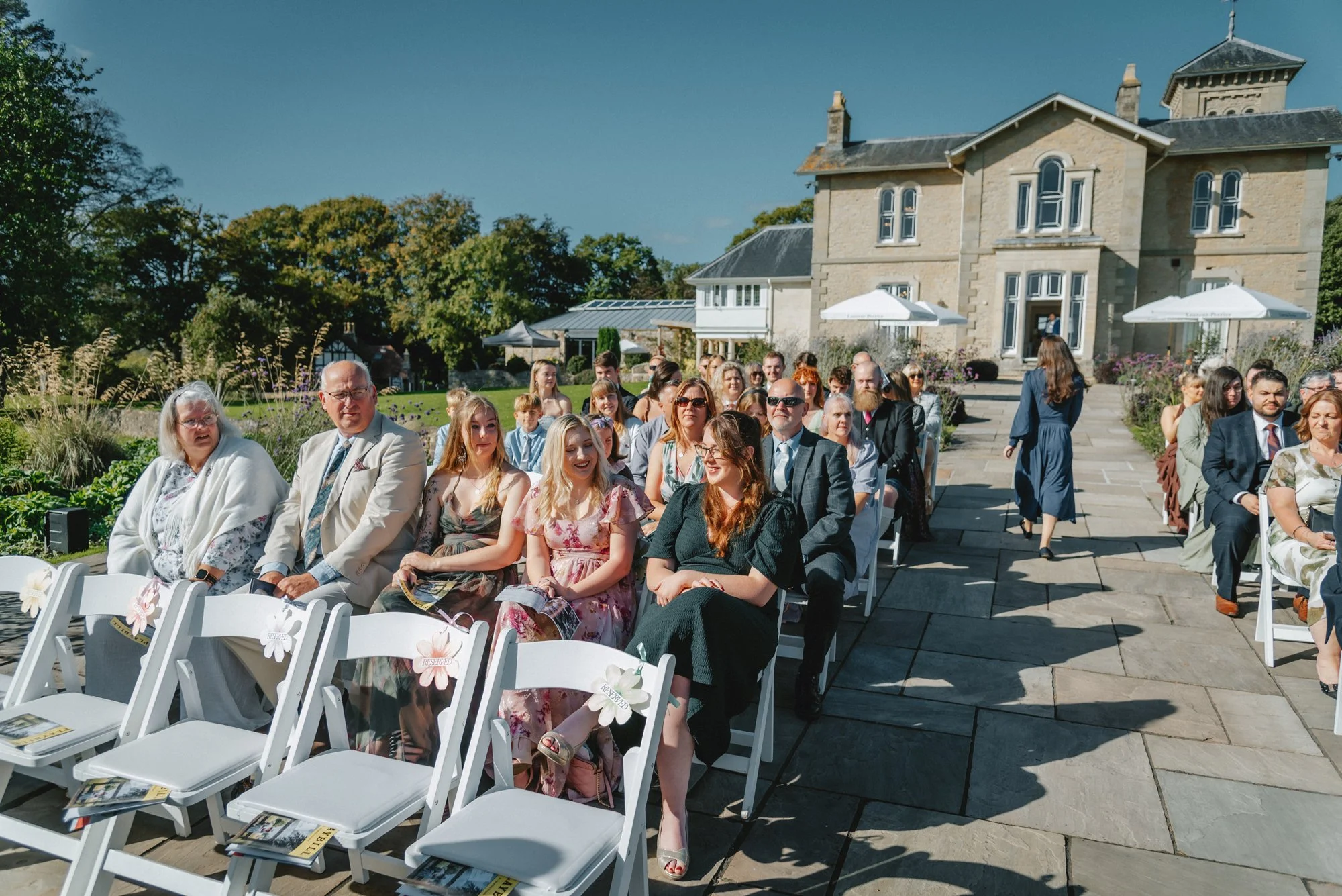 Guests seated outdoors at a wedding ceremony on a sunny day in front of a large historic house with patio umbrellas and greenery.