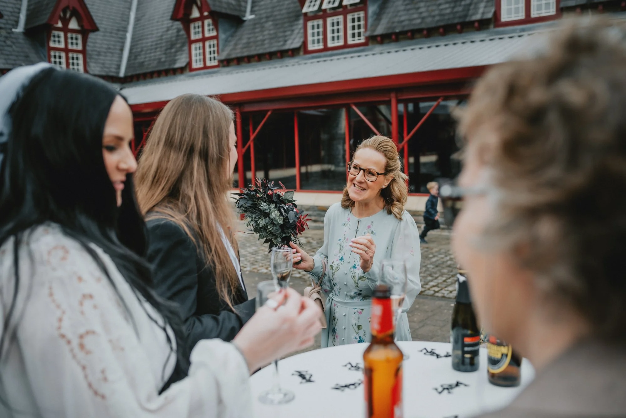 Four women conversing outdoors at a celebration, one holding a bouquet, some with drinks, with a restaurant or building with architectural details in the background.
