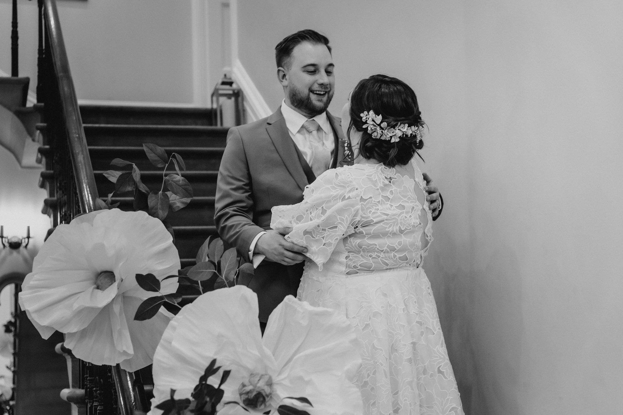 A black-and-white photo of a man in a suit holding a woman in a lace wedding dress and floral headband, standing on a staircase decorated with large paper flowers and leaves, sharing a joyful moment.