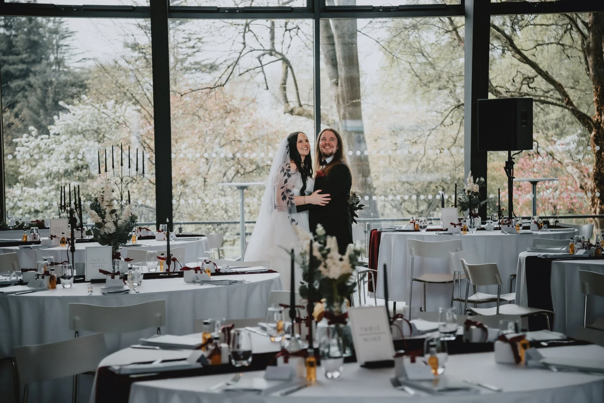 A bride and groom smiling and embracing inside a decorated reception hall with large floor-to-ceiling windows showing trees outside.