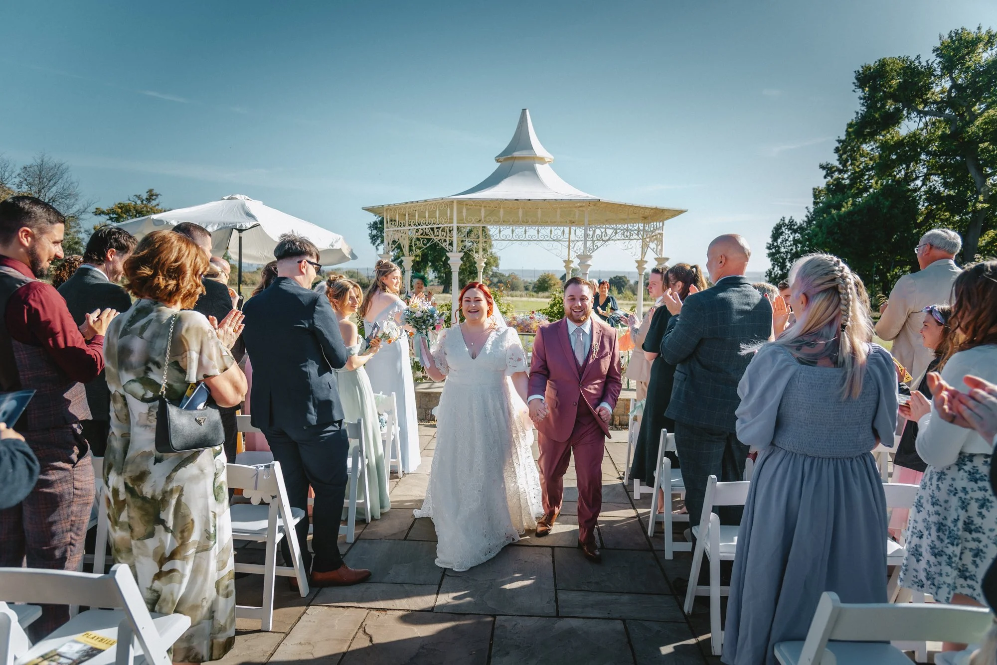 A newly married couple celebrating their wedding outdoors with guests clapping around them under a gazebo with white chairs and umbrellas on a sunny day.