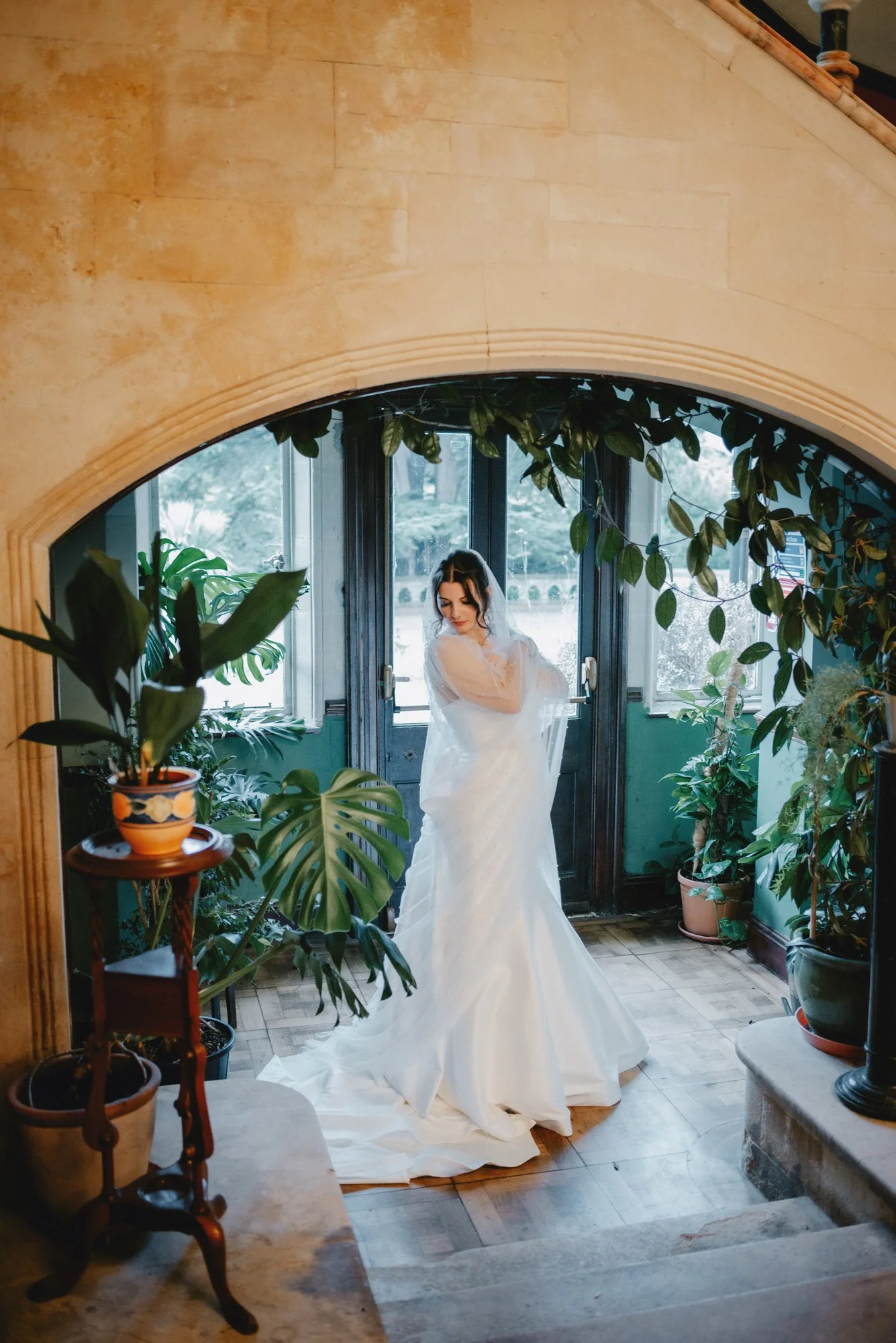 A bride in a white wedding dress and veil standing inside an arched doorway, surrounded by potted plants and natural light.