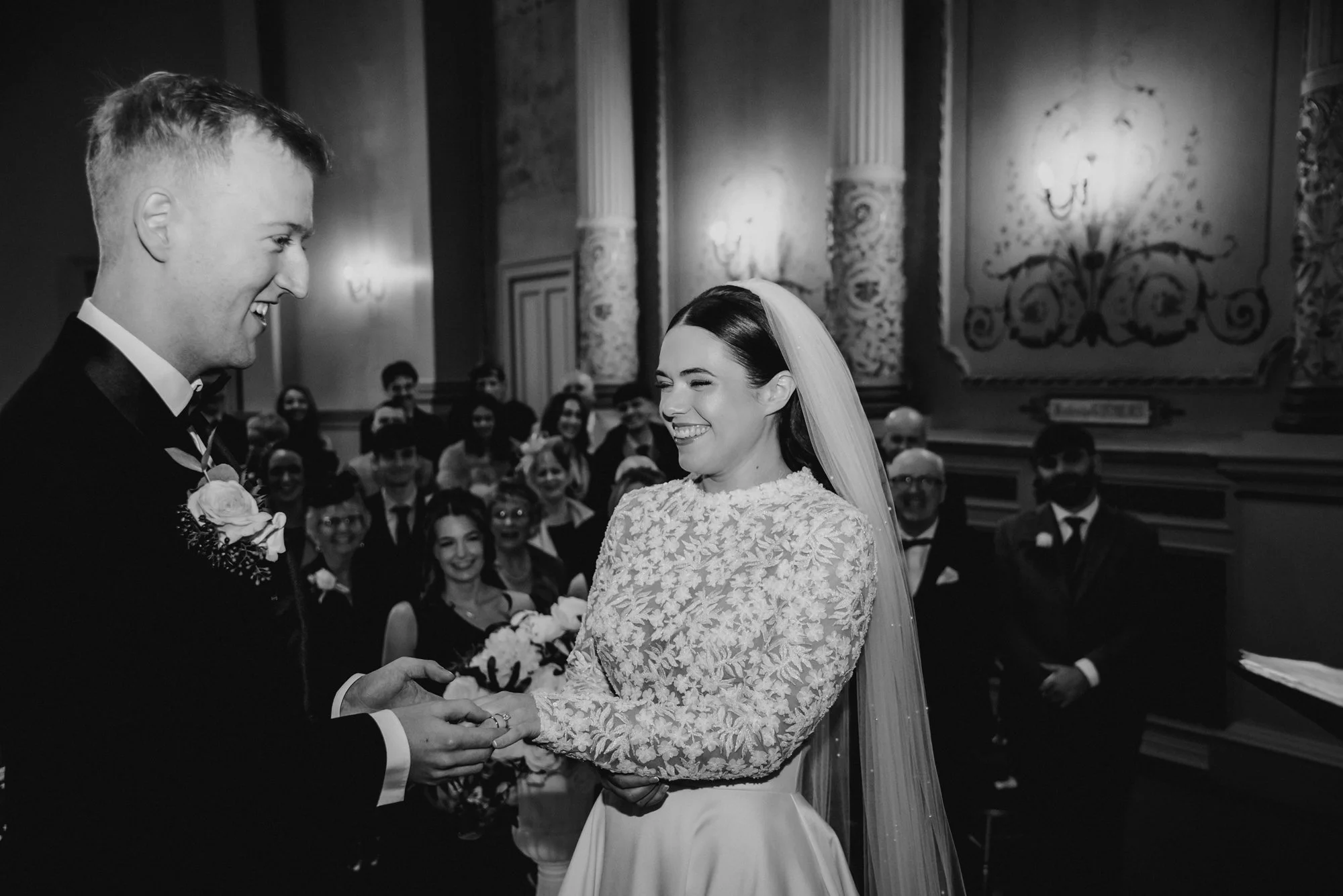 A black-and-white photo of a wedding ceremony showing a bride and groom exchanging rings, with smiling guests in the background.