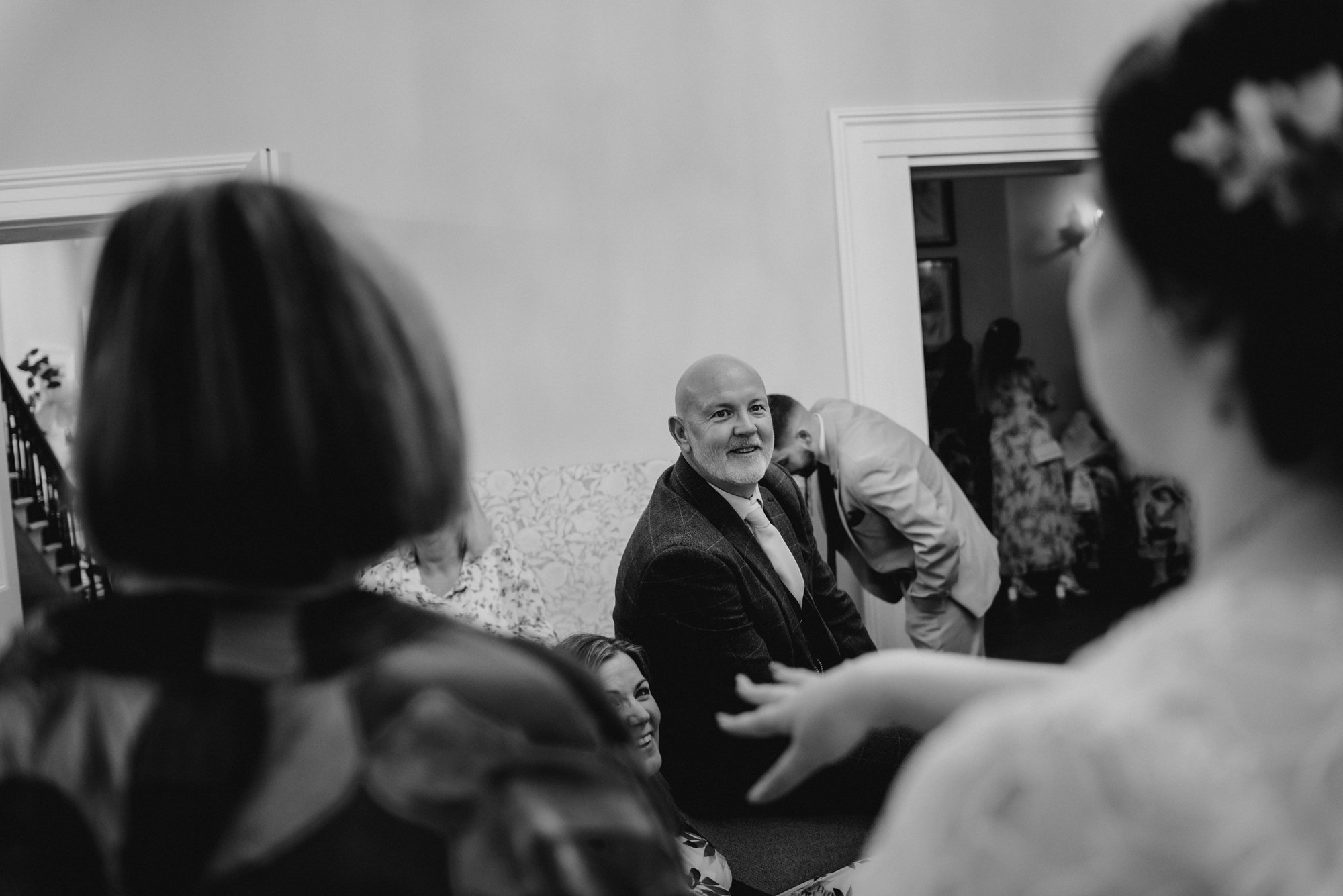 Black and white photo of a social gathering with several people, with one man wearing a suit and tie speaking or gesturing to others in a room.