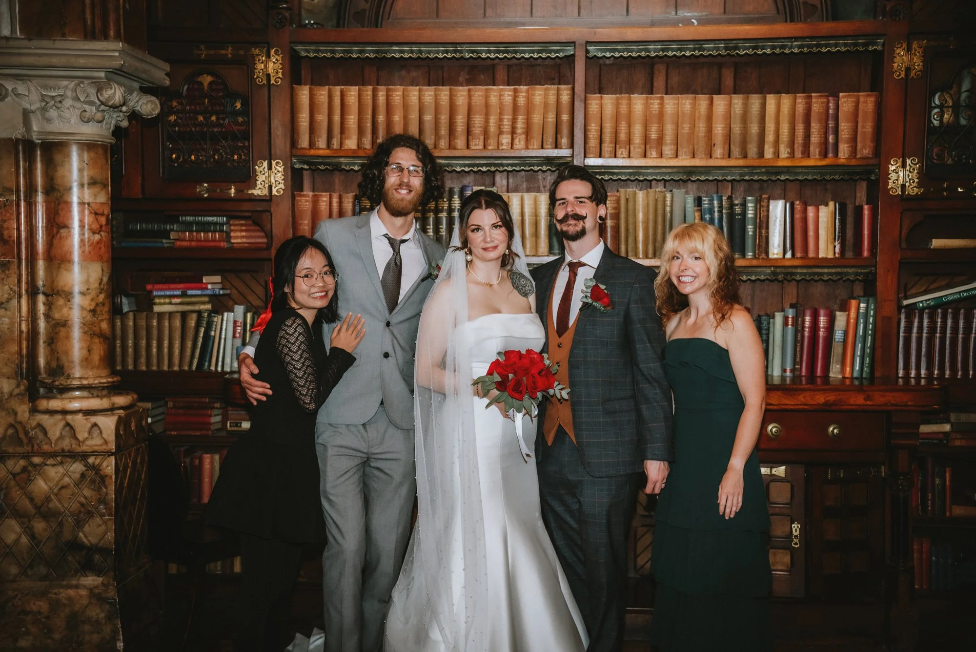 Group of six people at a wedding, including the bride in a white dress holding a bouquet of red roses, and a groom in a plaid suit with a red rose boutonniere, standing in front of a vintage wooden bookshelf filled with books.