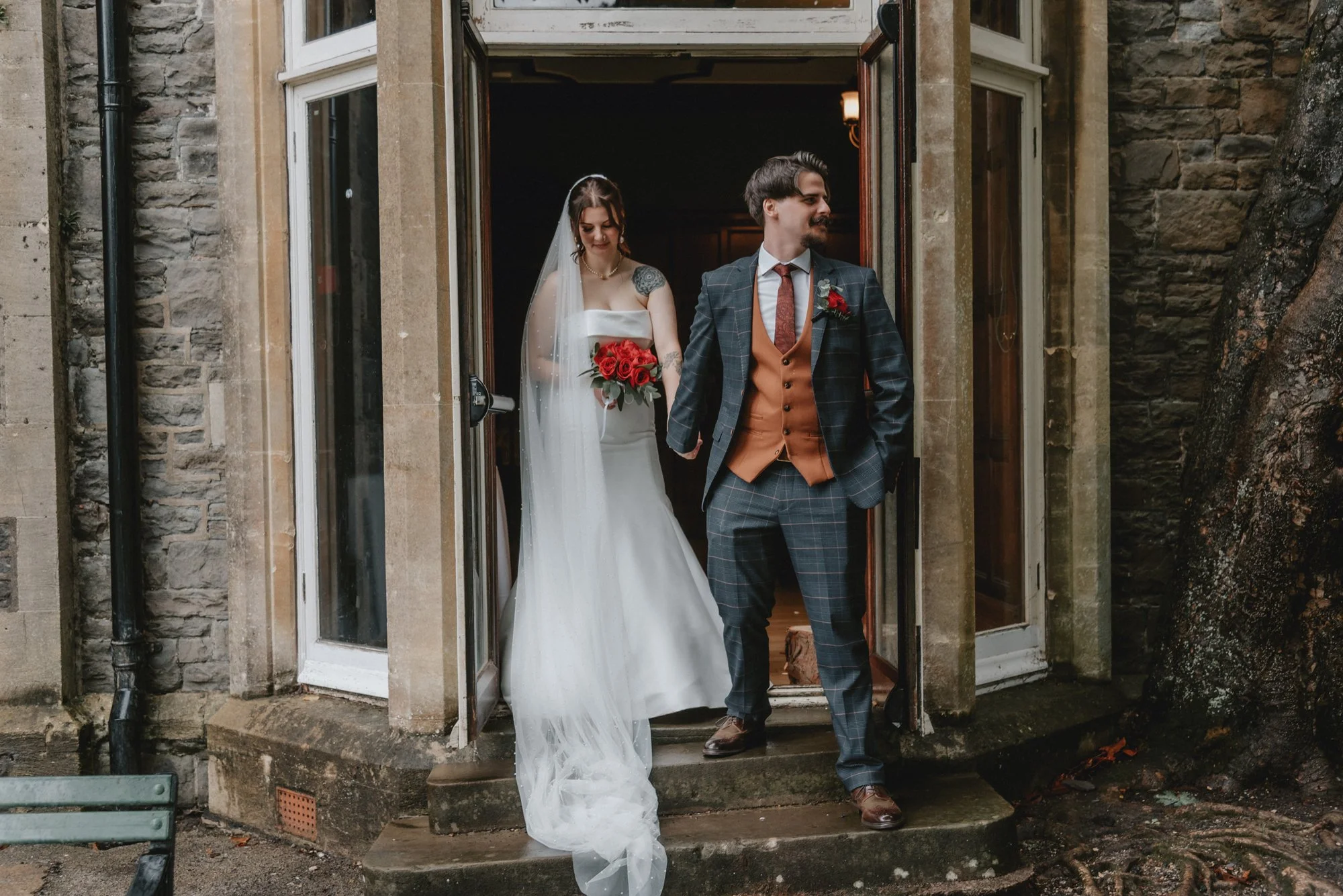 Bride in a white wedding dress and veil holding a bouquet of red roses, walking out of a building with her groom in a dark plaid suit with a tan vest and tie, holding her hand.