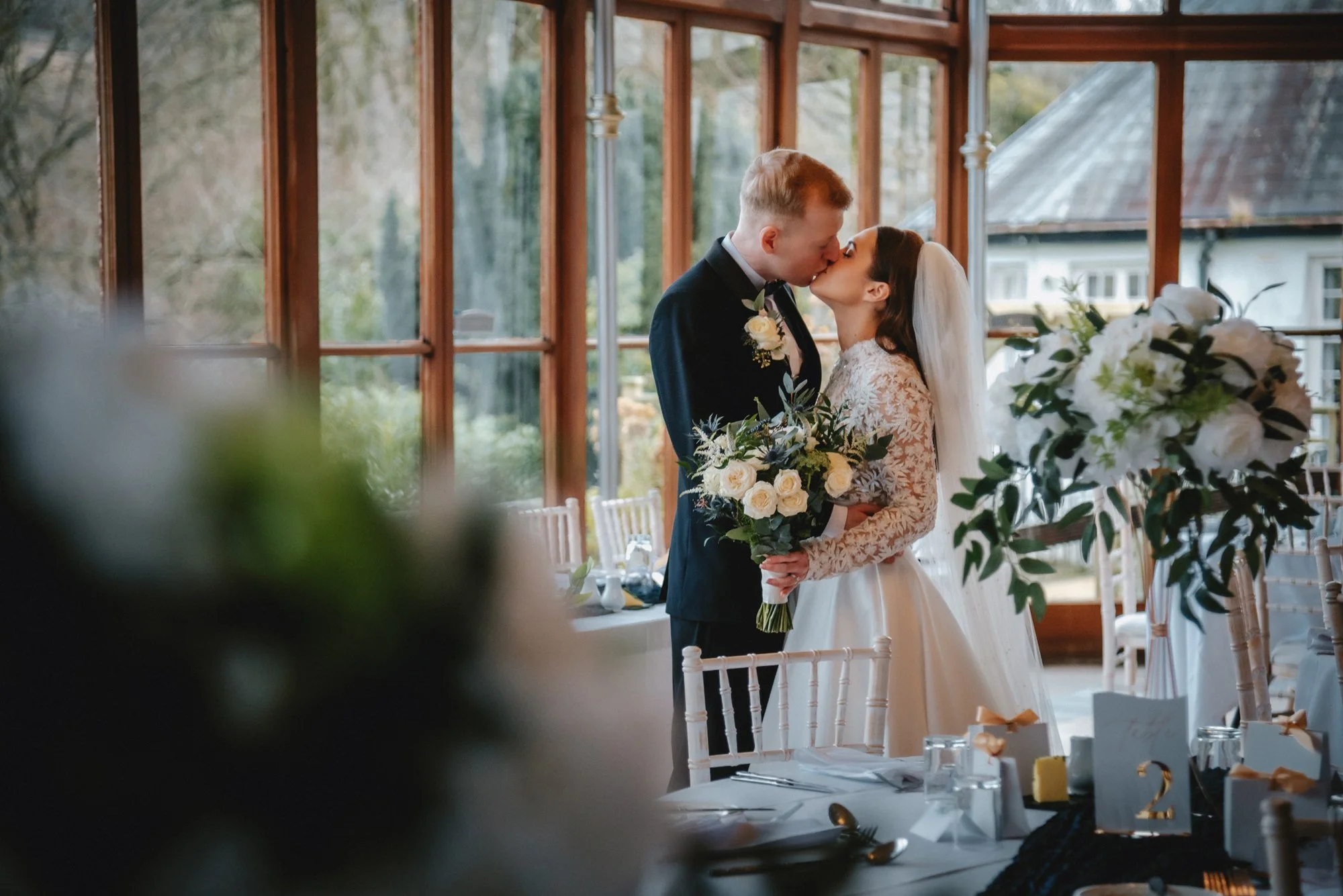A newlywed couple sharing a kiss indoors, with the bride holding a bouquet of white roses and greenery, and the groom in a black suit, near large windows and floral arrangements.