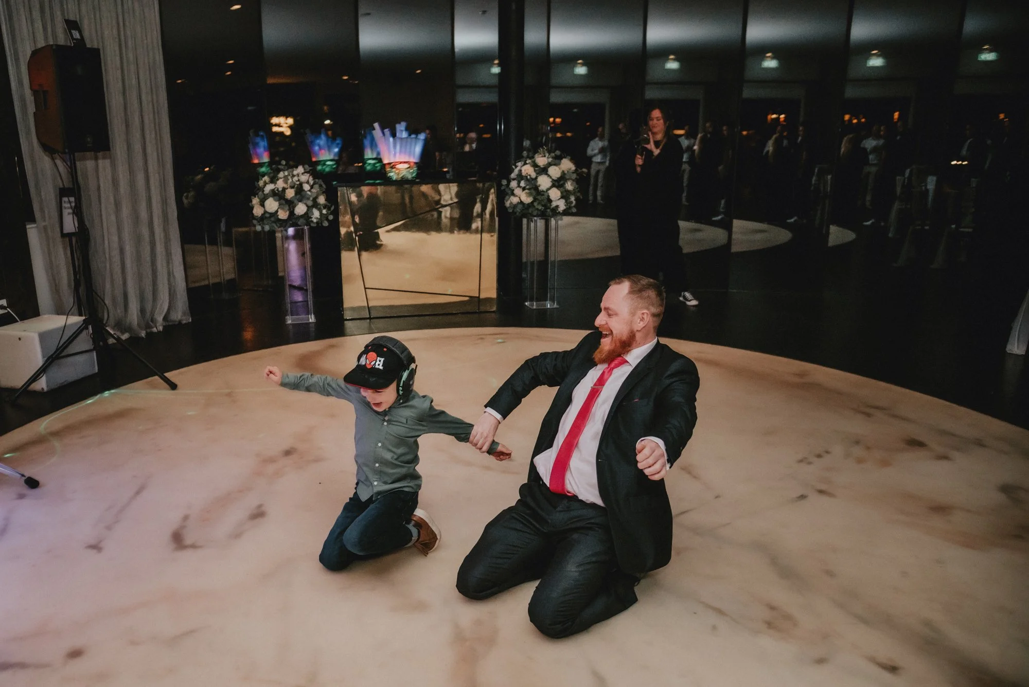 A man and a boy are dancing and having fun on a marble floor at an indoor event. The man, dressed in a suit with a red tie, is kneeling and smiling, holding the boy's hand. The boy is wearing a gray shirt, jeans, a hat, and large headphones, and is k